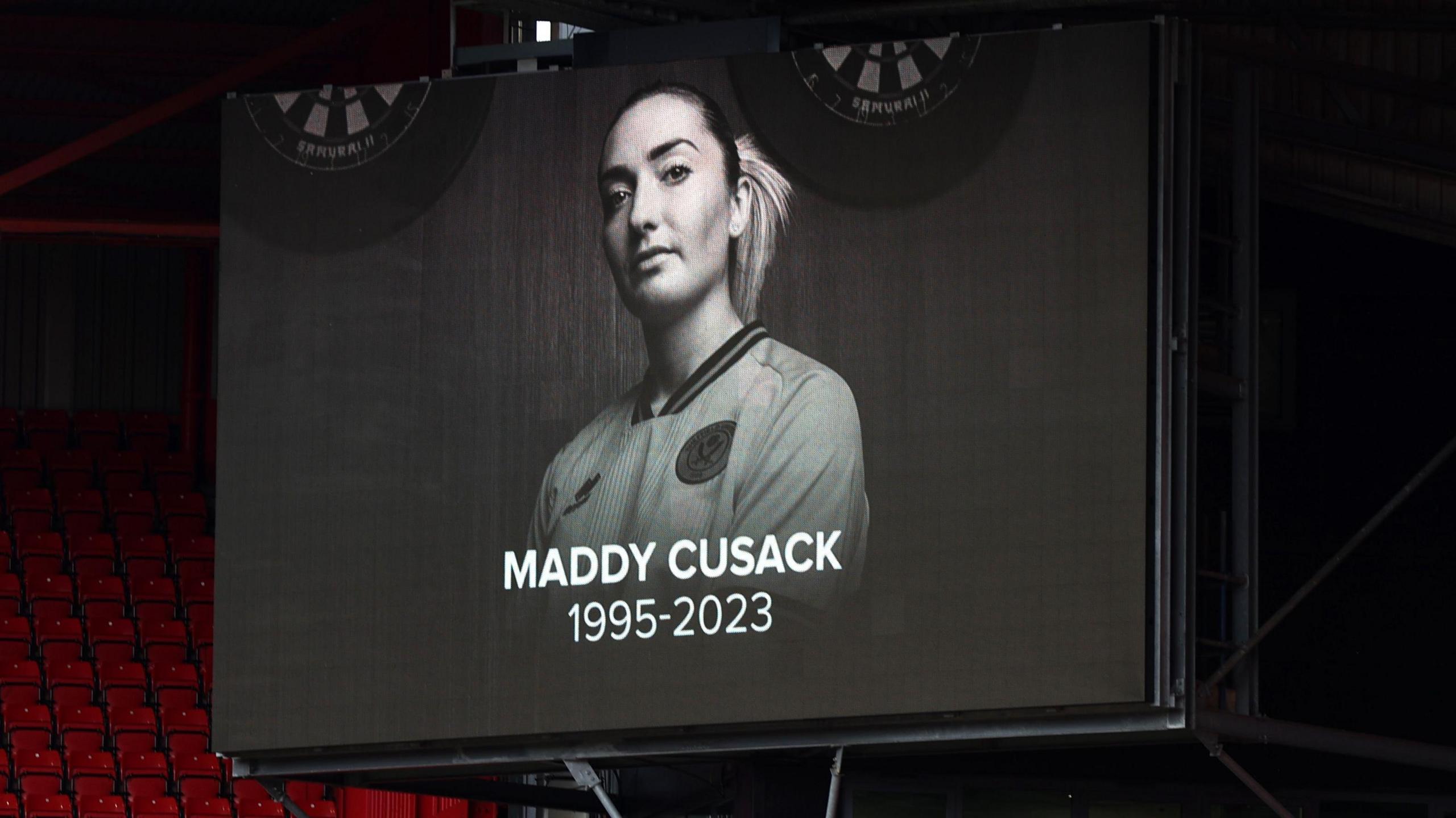 A screen displays a tribute in memory of former Sheffield United player Maddy Cusack during a match between Sheffield United and Crystal Palace. It's a black and white photo of Maddy in her kit, her hair tied back in a pony tail. Beneath it reads: Maddy Cusack 1995-2023.