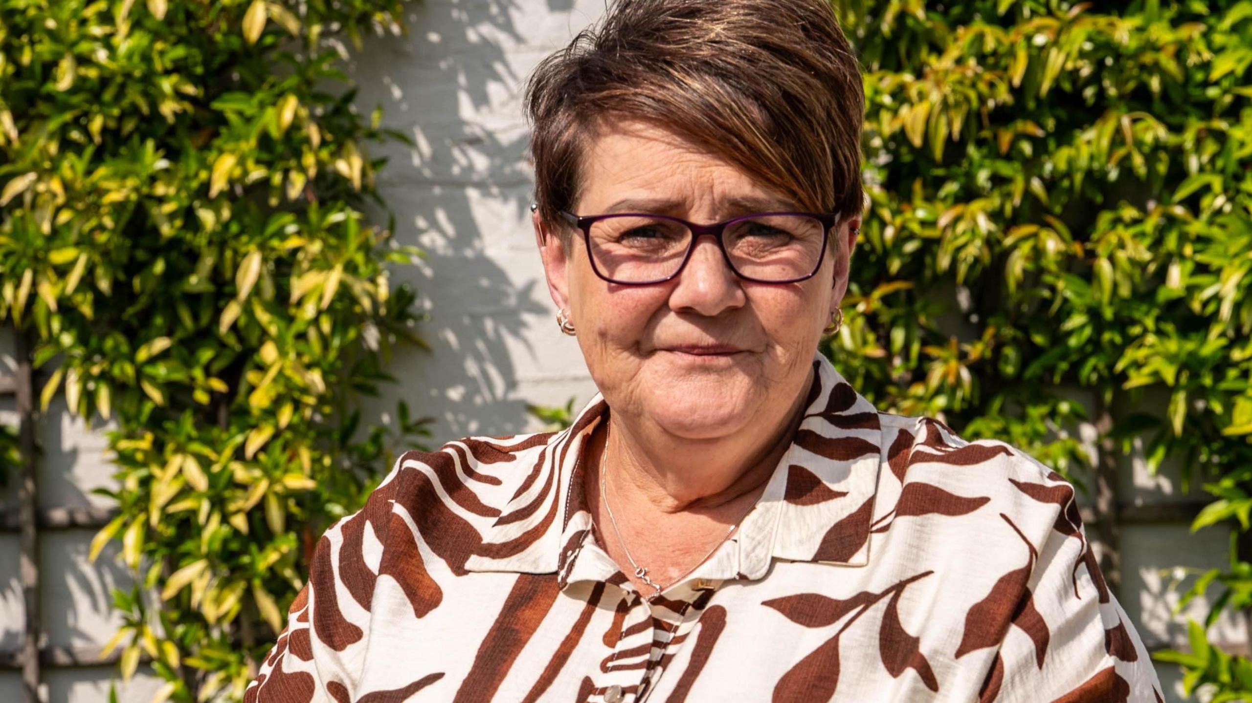 A head and shoulders picture of Tracy Page, wearing a brown and cream patterned top, sitting in front of a wall with climbing plants.