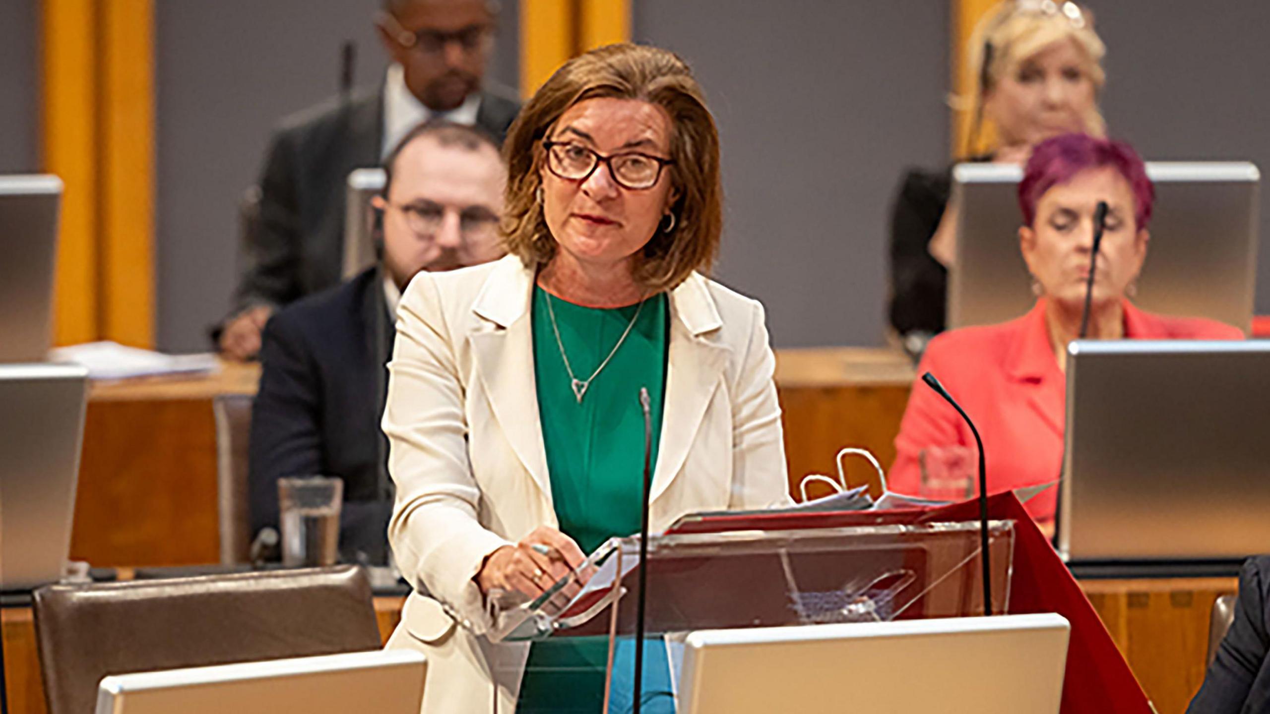 Eluned Morgan stood addressing the Senedd, in a white jacket and green dress.