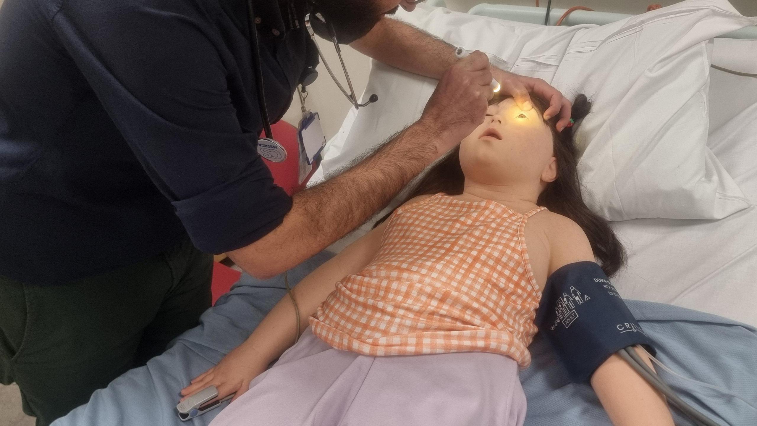 A hospital worker shining a light into the manikin's eye with a torch as it lays on a bed.