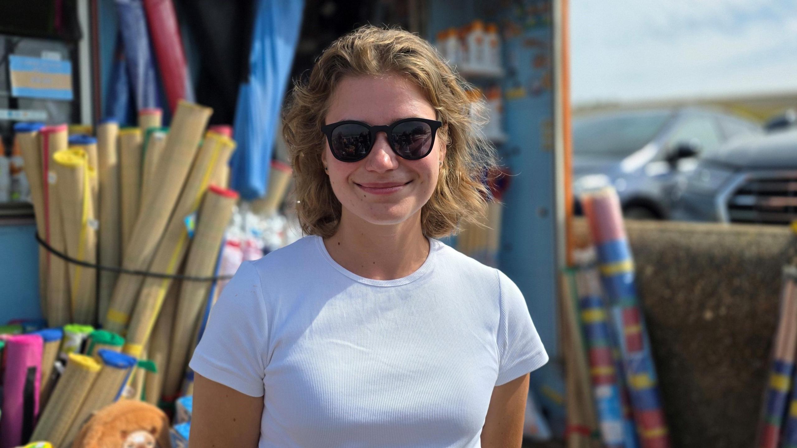 We can see a young woman in a white t-shirt wearing shades. Behind her is a stall selling various goods for the beach - like wind-breakers.