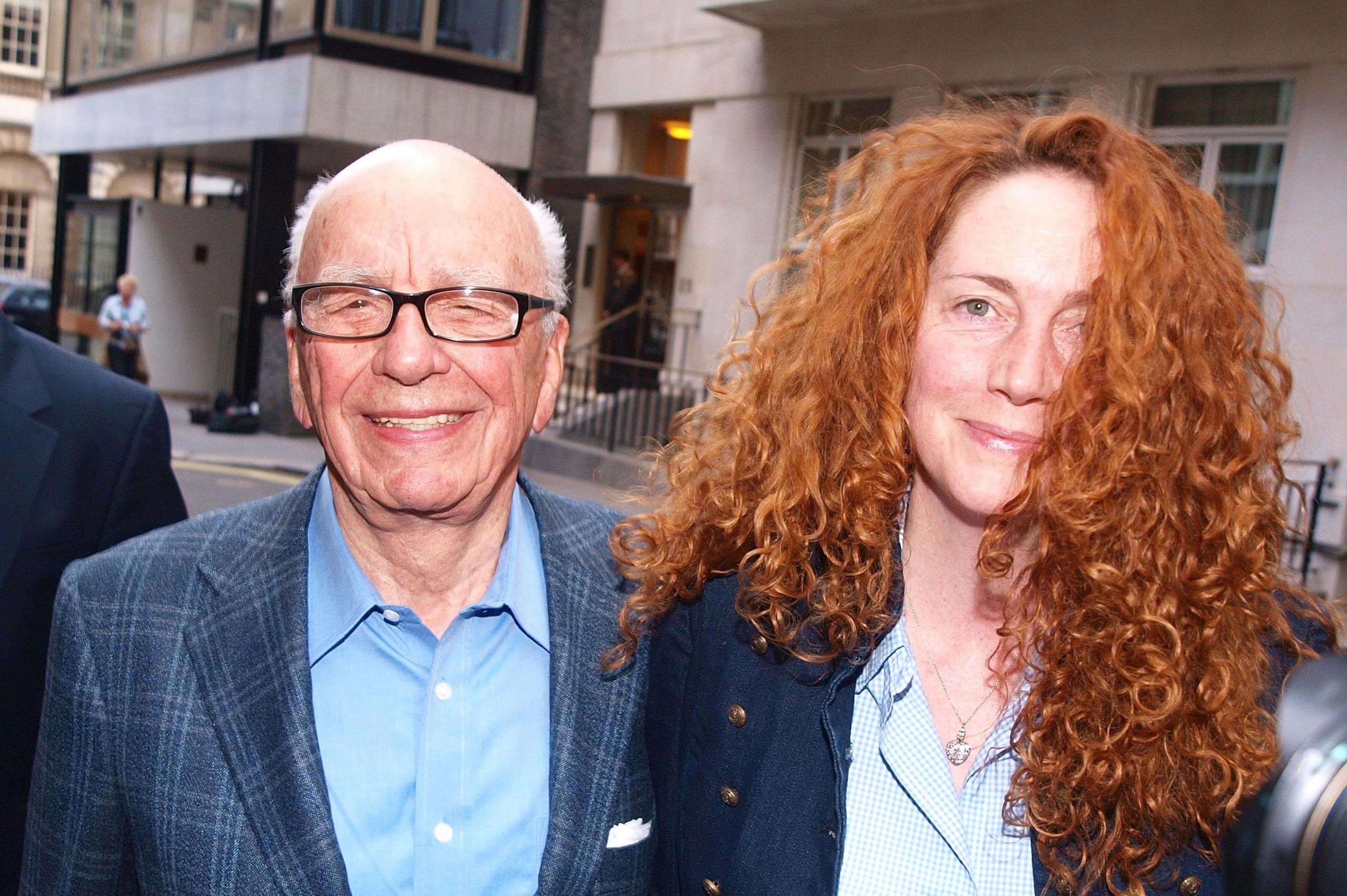 Rupert Murdoch and Rebekah Brooks smile at the camera. Rupert Murdoch has short grey hair and has glasses. He wears a blue checked jacket and pale blue shirt. Rebekah Brooks is wearing a blue jacket and checked shirt. She has long wavy read hair.