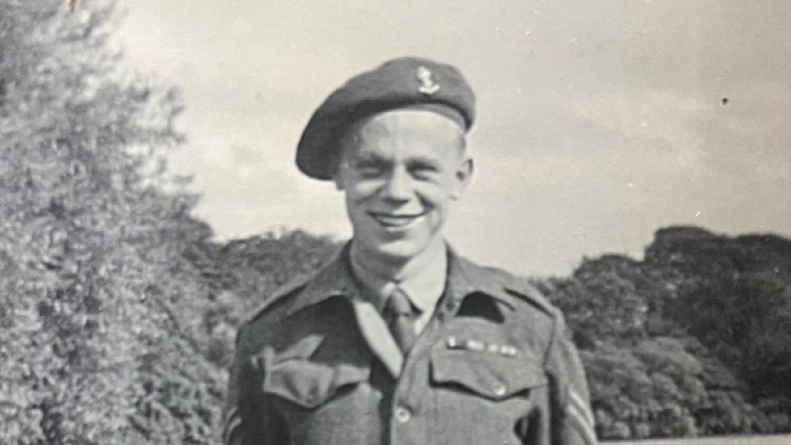 Black and white picture of Stanley Eastgate in his army uniform during the Second World War, he has a beret on his head and his hands behind his back, he is in a field with trees visible in the background and a small child stands looking on on the right hand side.