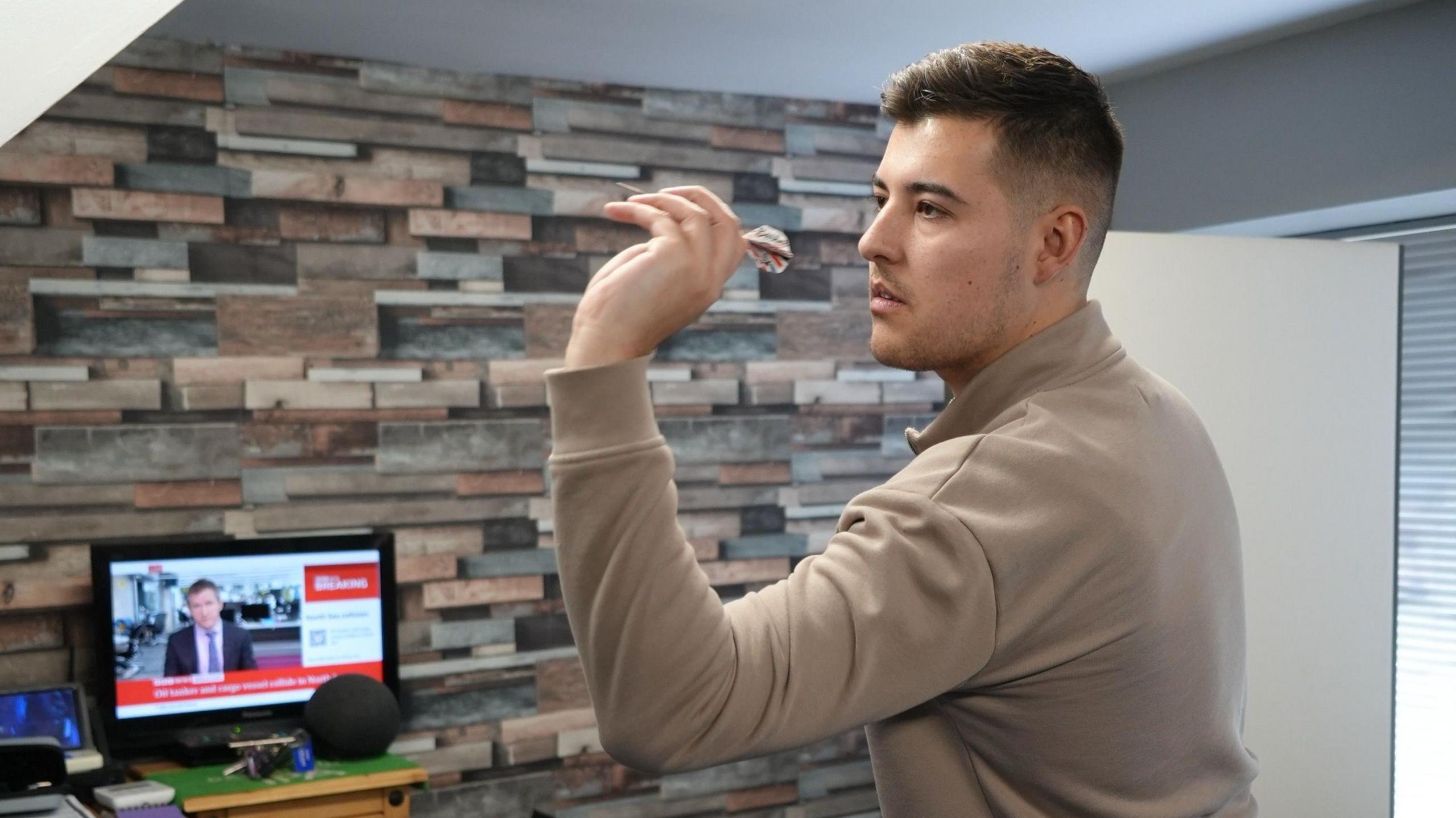 Meikle is pictured aiming a dart at a dartboard. A TV can be seen behind him with BBC News on. 