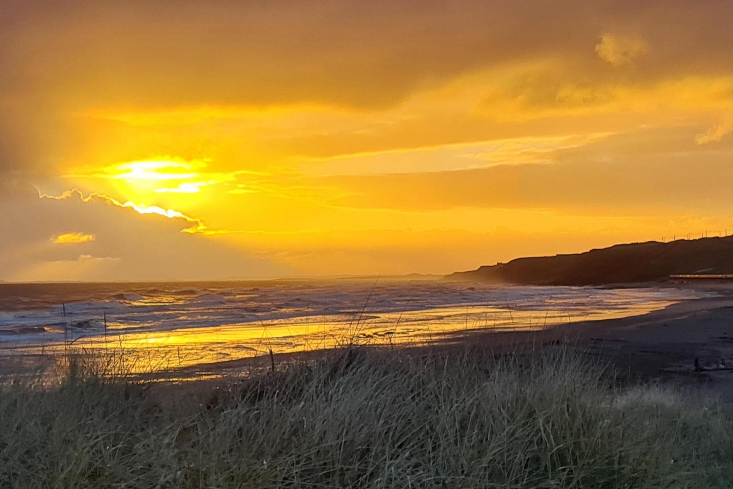 Rays of light break through the clouds above the coast. Below, waves crash onto the sandy beach, which is bordered by dunes.