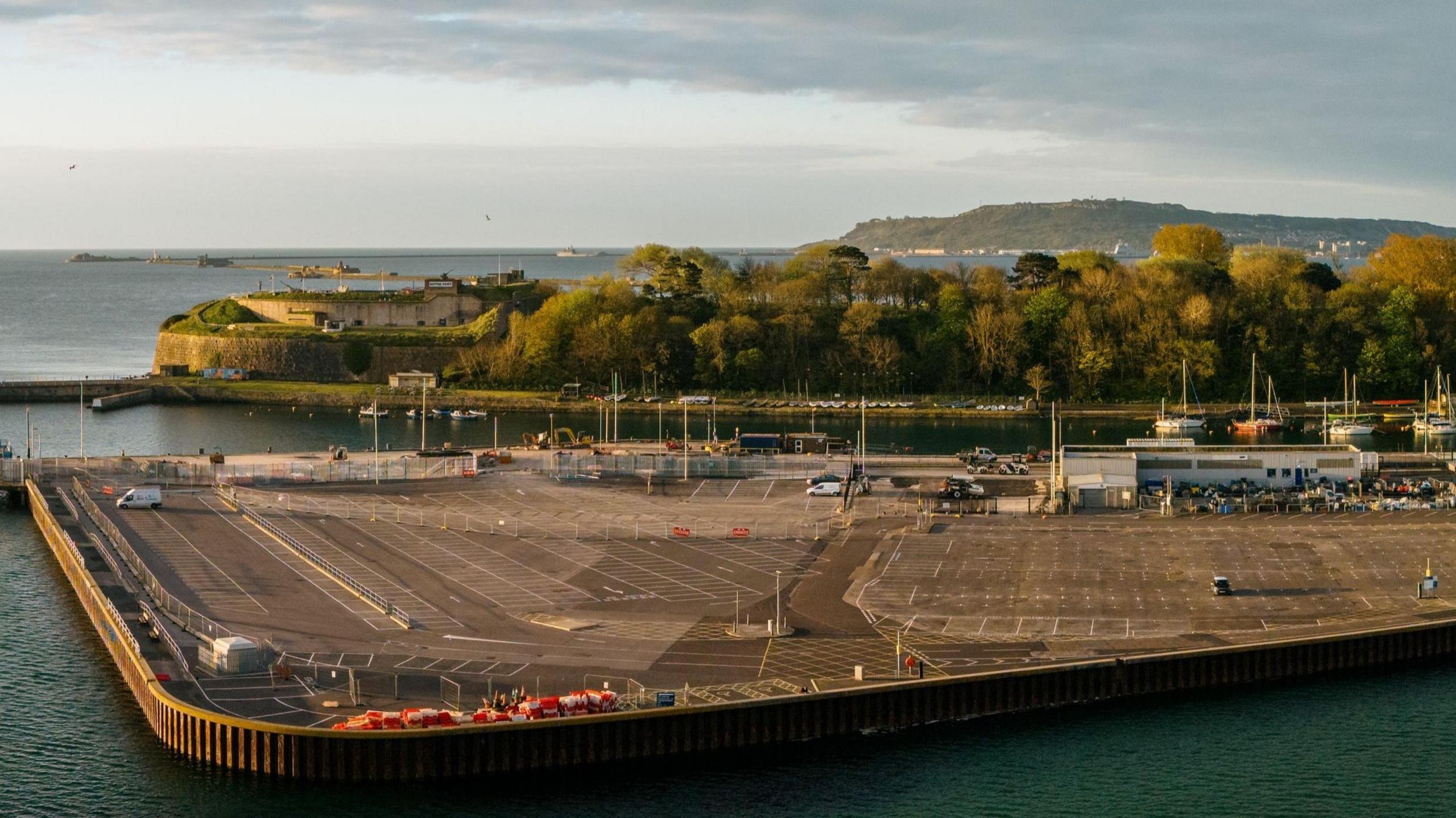 A drone photo of a large expanse of land jutting out into the sea. There's a car park and some buildings on it.