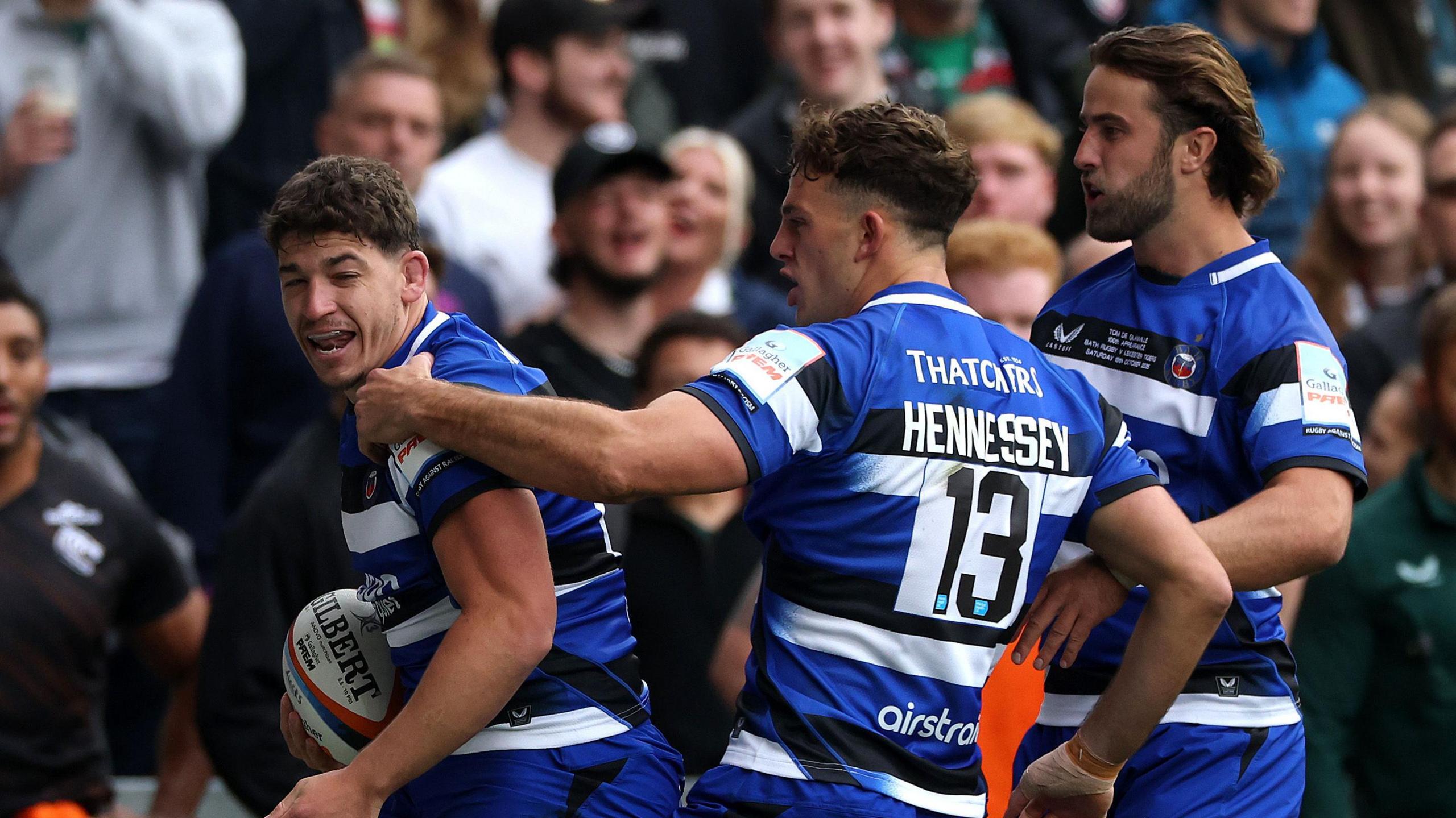 Three Bath Rugby players, all wearing the blue, black and white strip celebrate following Dan Frost's try. Tom De Glanville and Louis Hennessey prepare to hug Frost with fans reacting in the background.