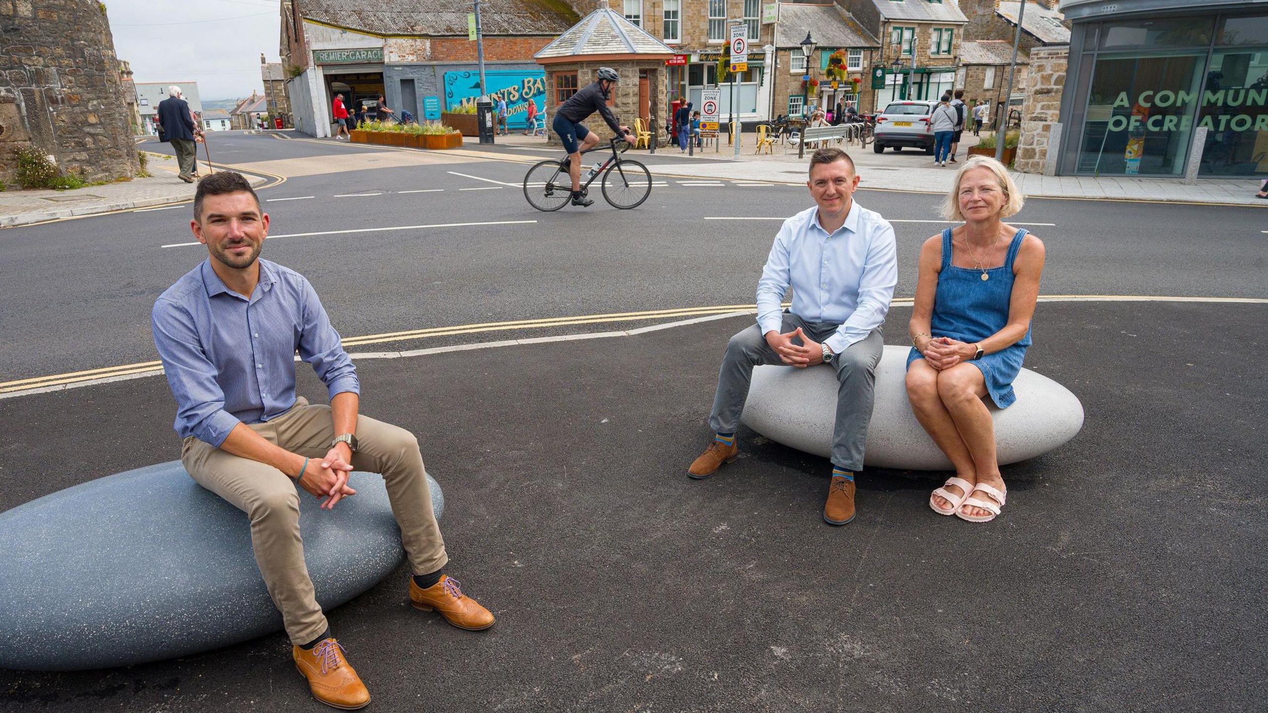 One man is sitting on a giant pebble which sits on tarmac while a man and a woman share another one. They have their hands folded and are smiling at the camera. Behind them is a road with a cyclist on it and pedestrians on the adjacent pavements walking past shops.