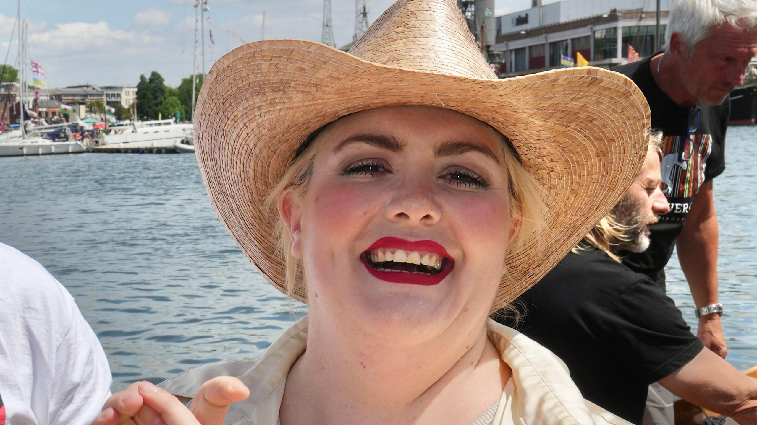 Artist Jayde Adams smiling at the camera wit hredlipstick on and blonde hair, wearing a straw hat, on a boat in the Bristol Harbour