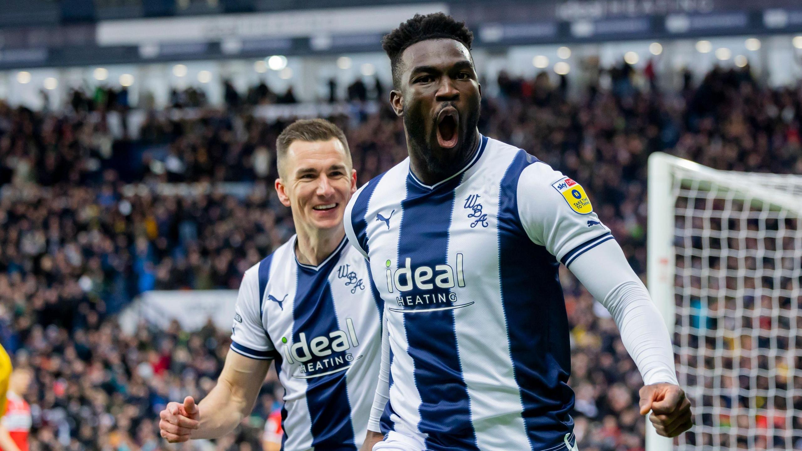 Daryl Dike celebrates scoring for West Bromwich Albion. He is wearing a dark blue and white striped home shirt and is bellowing out as he starts his celebratory run. He is being chased by a smiling team-mate.