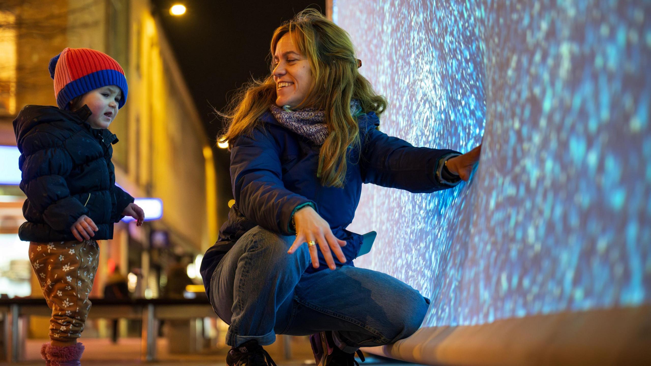 A woman pushes her hand into a large, flexible screen with light and dark sections. She is crouched down and smiling at her young son, who is standing next to her