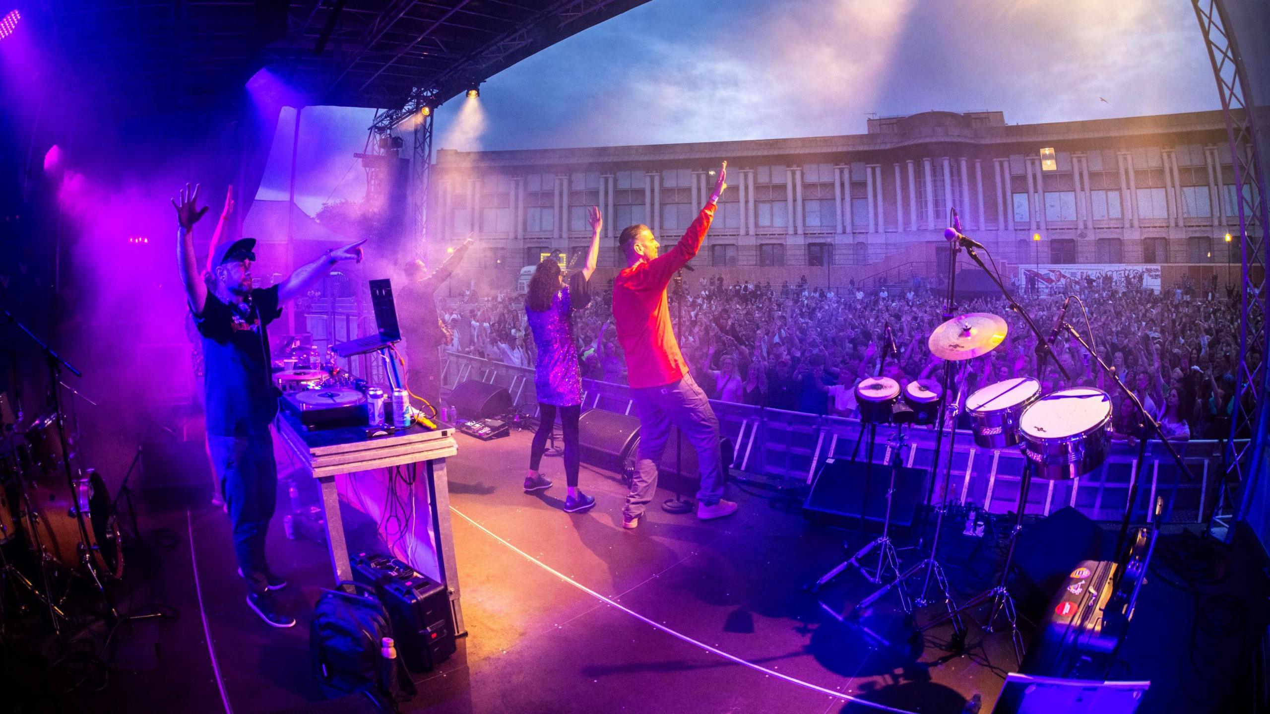 A band performs on stage at the Bristol Harbour Festival. The image is taken from the back of the stage looking out over the Lloyds Ampitheatre with a large crowd in the background. The stage and audience are illuminated by blue and purple lights