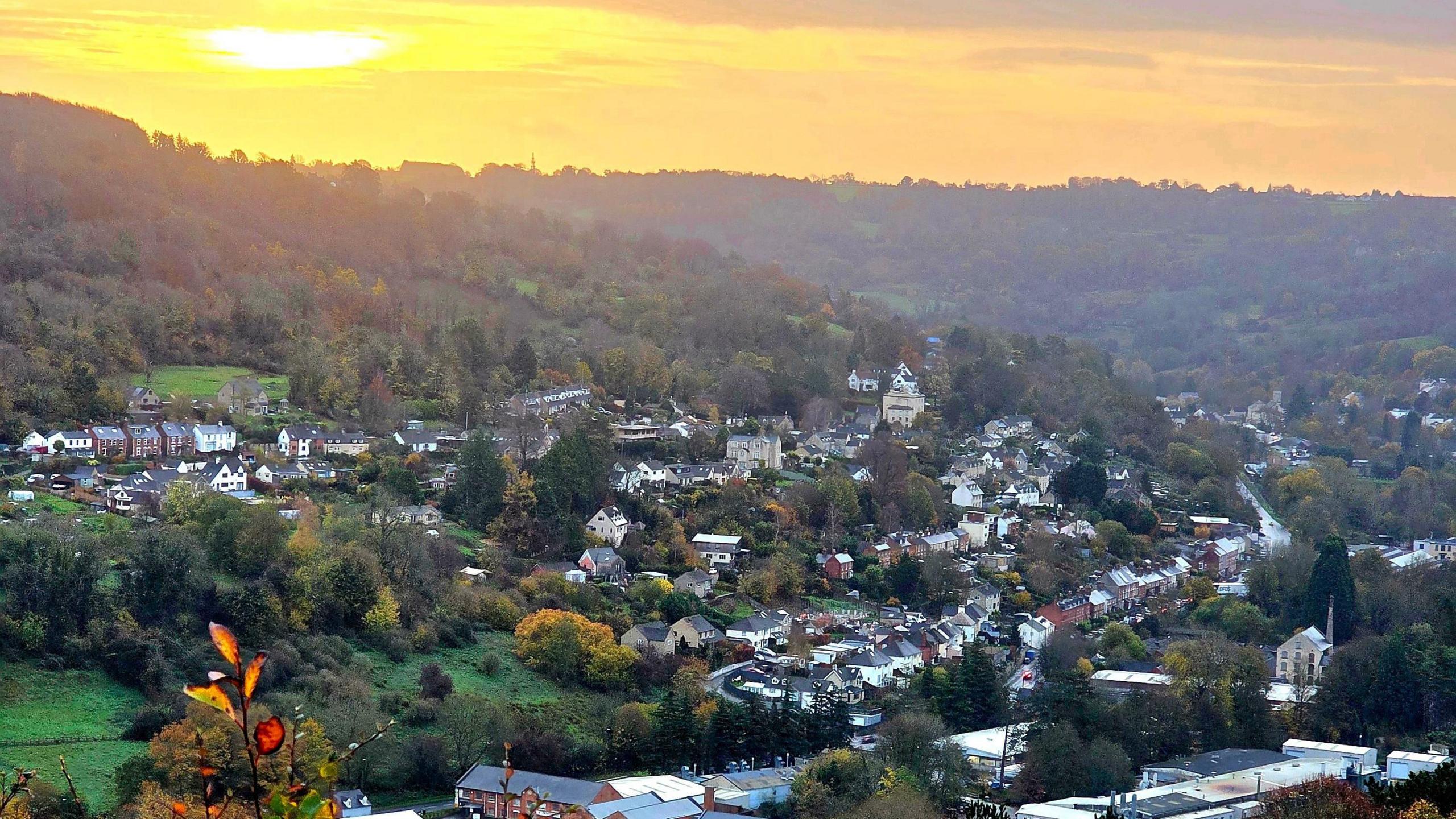 The sun rises above the wooded Stroud Valleys, painting the sky a golden hue. There are lots of houses in the valley, along with a major road. The trees are different shades of green, yellow, red and brown.
