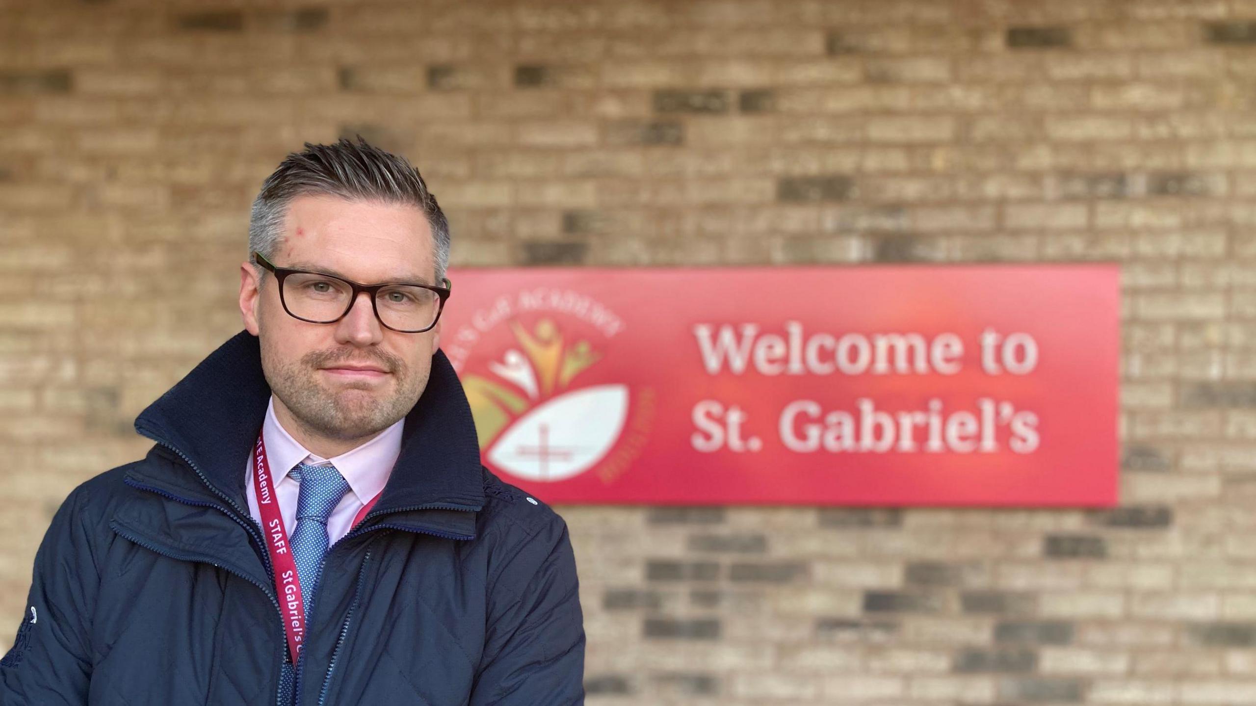 Headteacher Andy Taylor in front of a red sign on a sandy brick building that says Welcome to St. Gabriel's