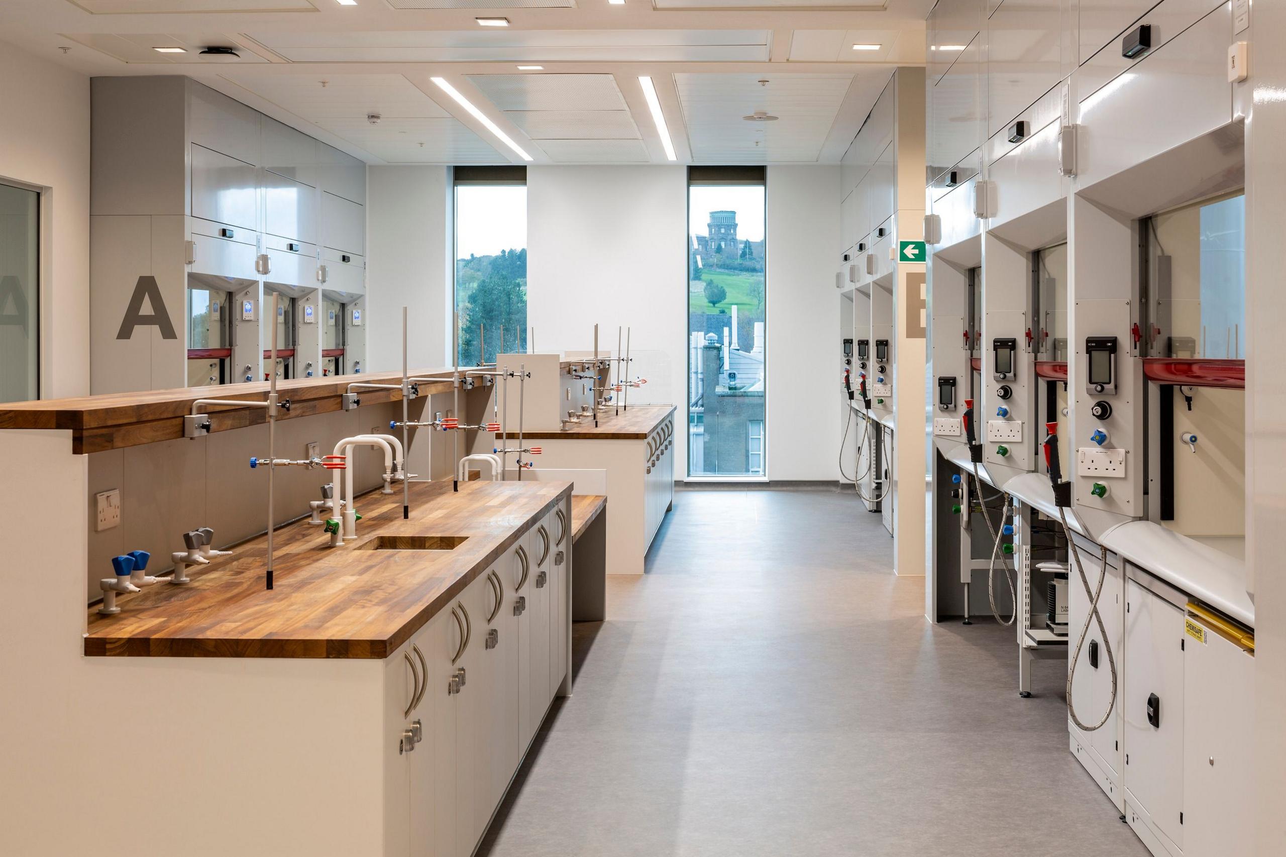 A laboratory area in The Nucleus - with light cupboard units, warm wooden worktops, scientific work stations down one side and tall windows showing a view of Calton Hill beyond.