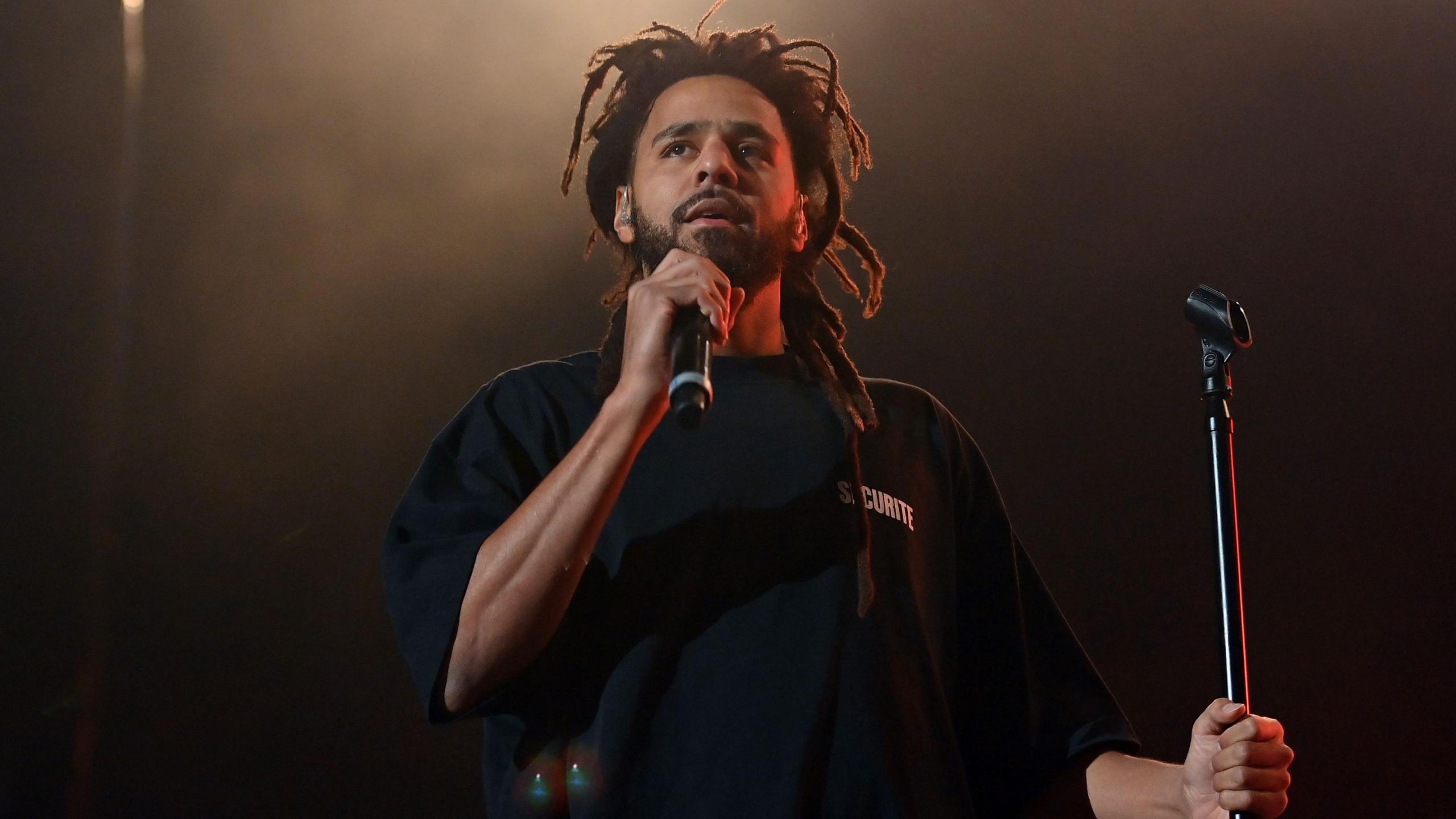 J Cole on stage. J Cole holds has dreads and a short beard. In his right hand, he holds a mic to his face, holding the mic stand with his left. 