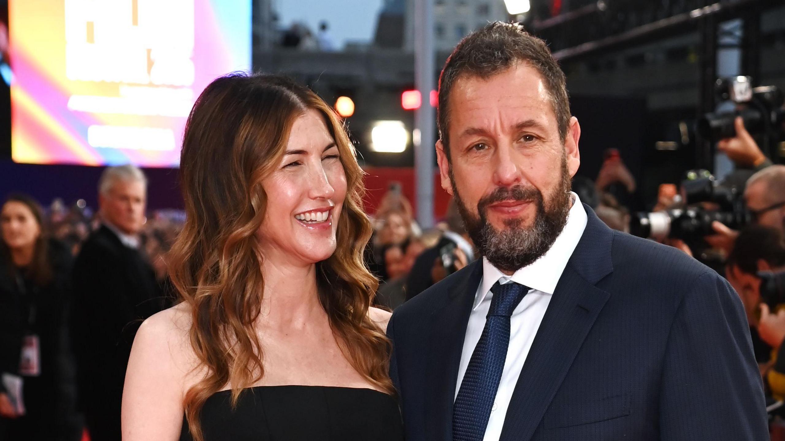 Jackie Sandler smiling at her husband Adam Sandler, as he poses on the red carpet