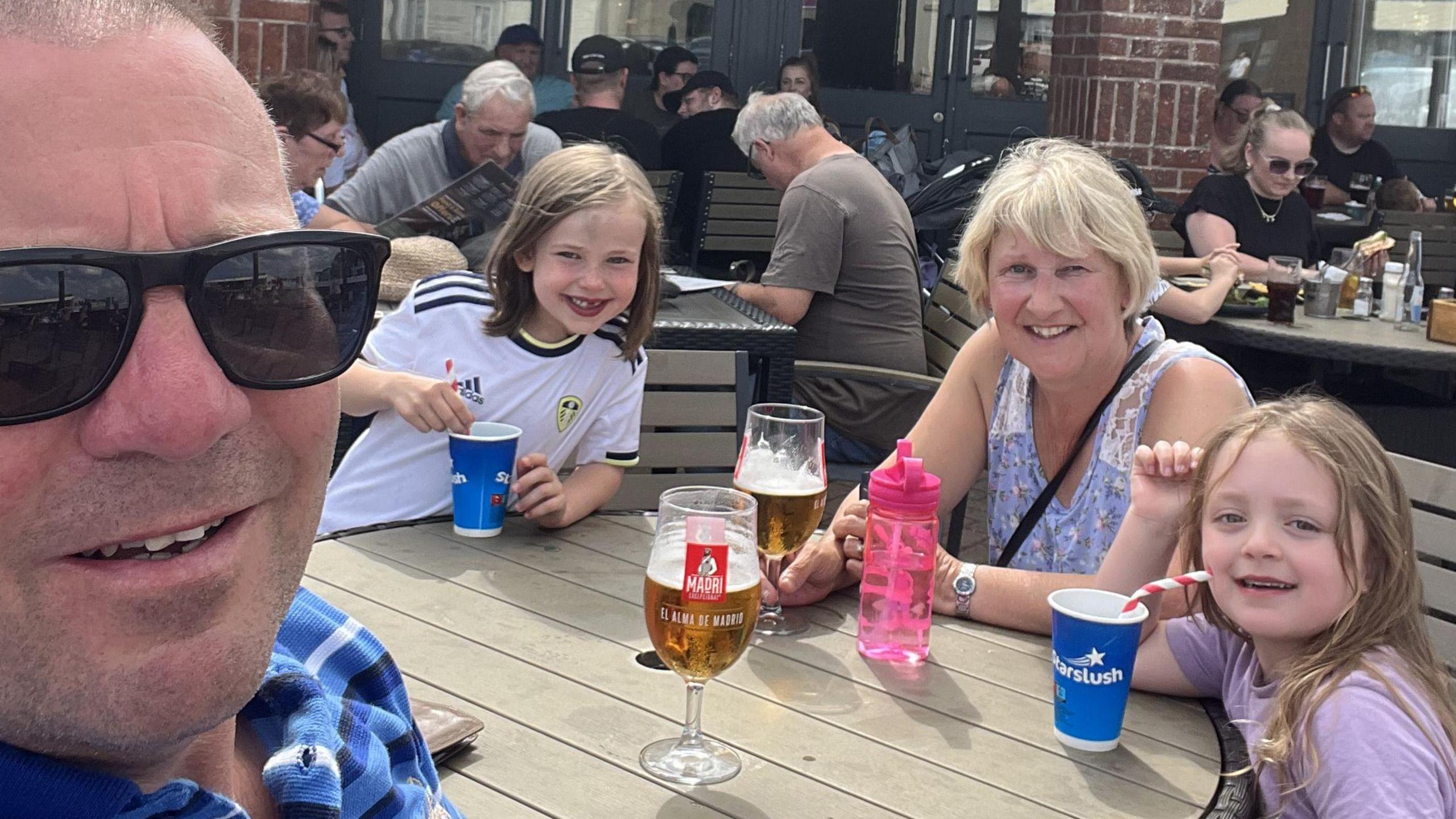 Andrew Dawson smiles close to the camera as he takes a selfie with his wife  Sue and two granddaughters at Skipsea Sands caravan park. They are sitting around a table in a beer garden, with pints of continental beer and slushy drinks for the children.