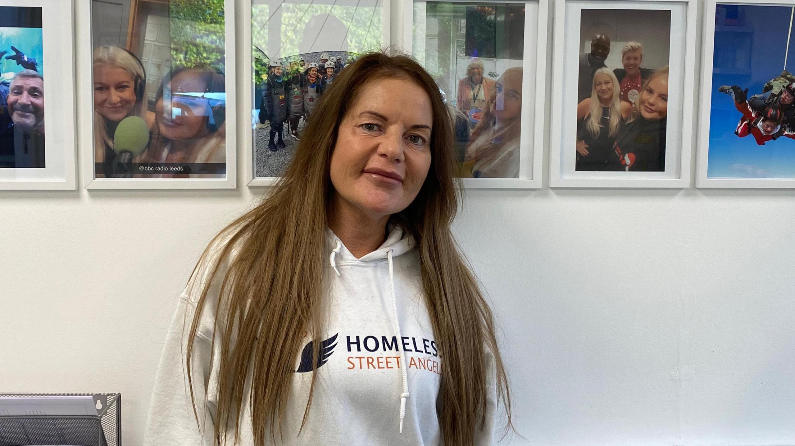 A woman with long brown hair stands in front of a gallery wall. She wears a hoodie with Homeless Street Angels emblazoned over the front