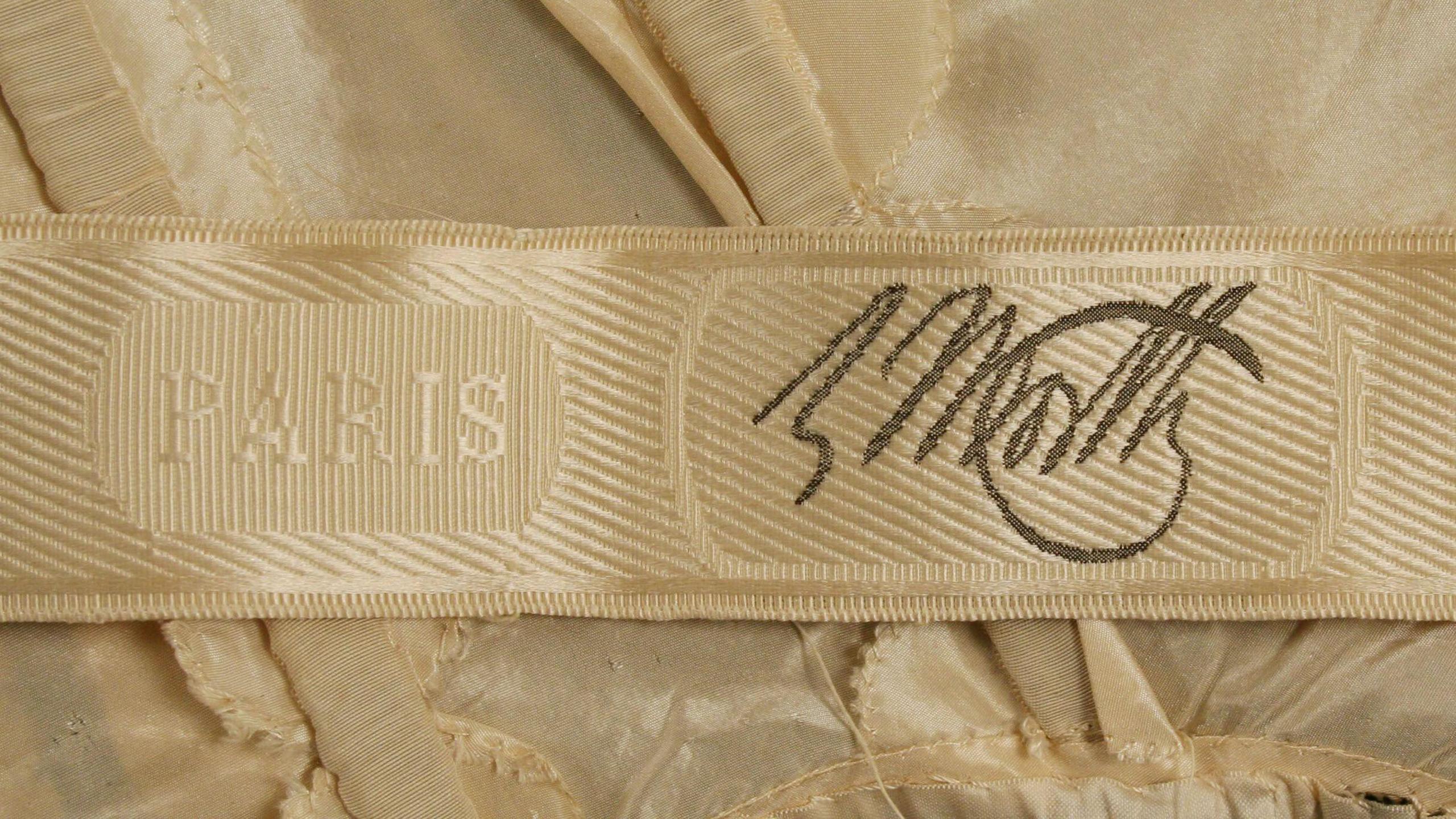 A close-up of a woven, golden-coloured Worth clothing label, which matches a golden-coloured dress. It is marked "Paris" in capital letters, followed by a Worth signature in dark grey-coloured script.