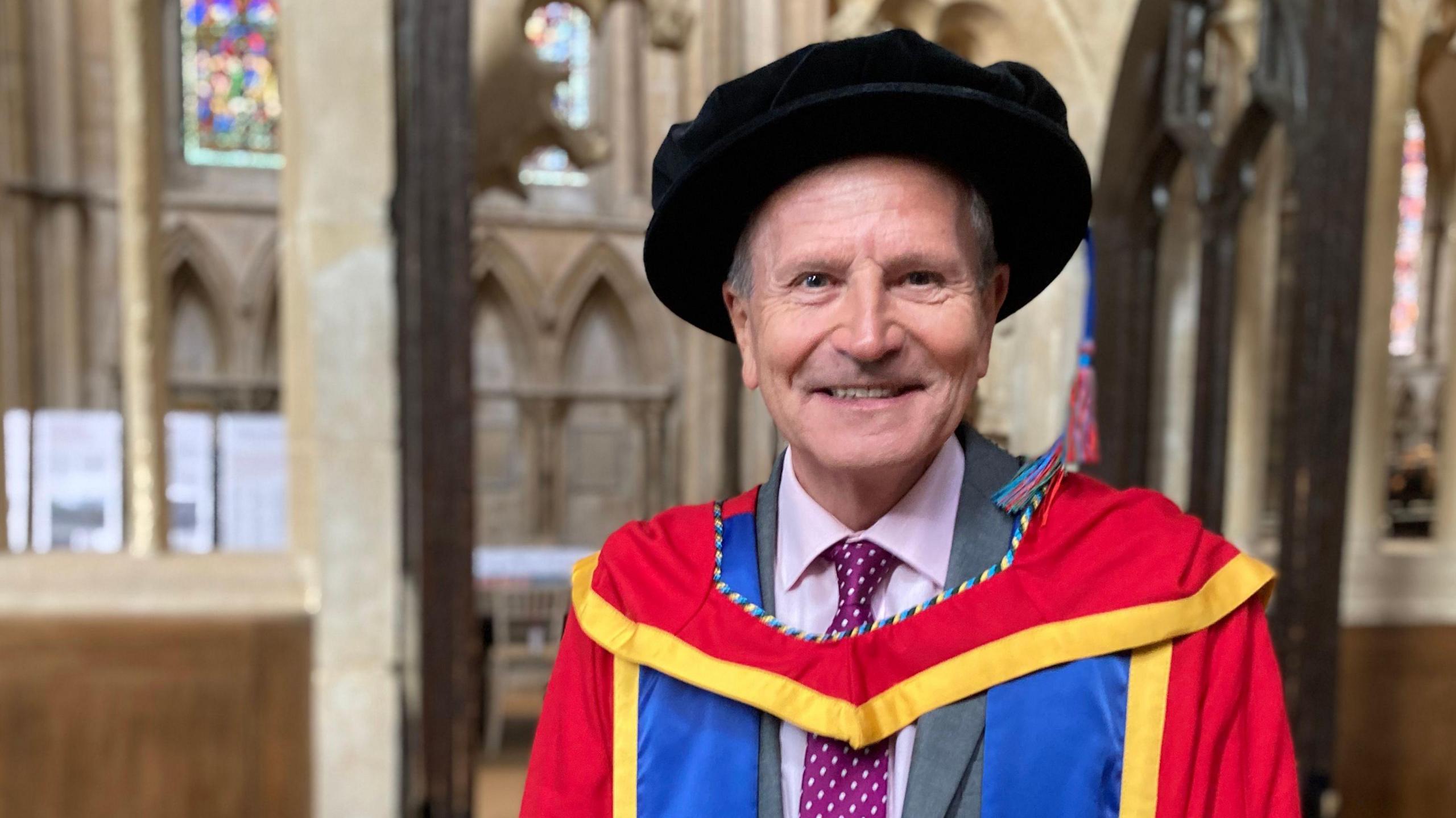 Peter Levy inside Lincoln Cathedral. He wears a black graduation hat and the red, blue and yellow robes of Bishop Grosseteste University over a grey suit and purple, polka-dot tie. 