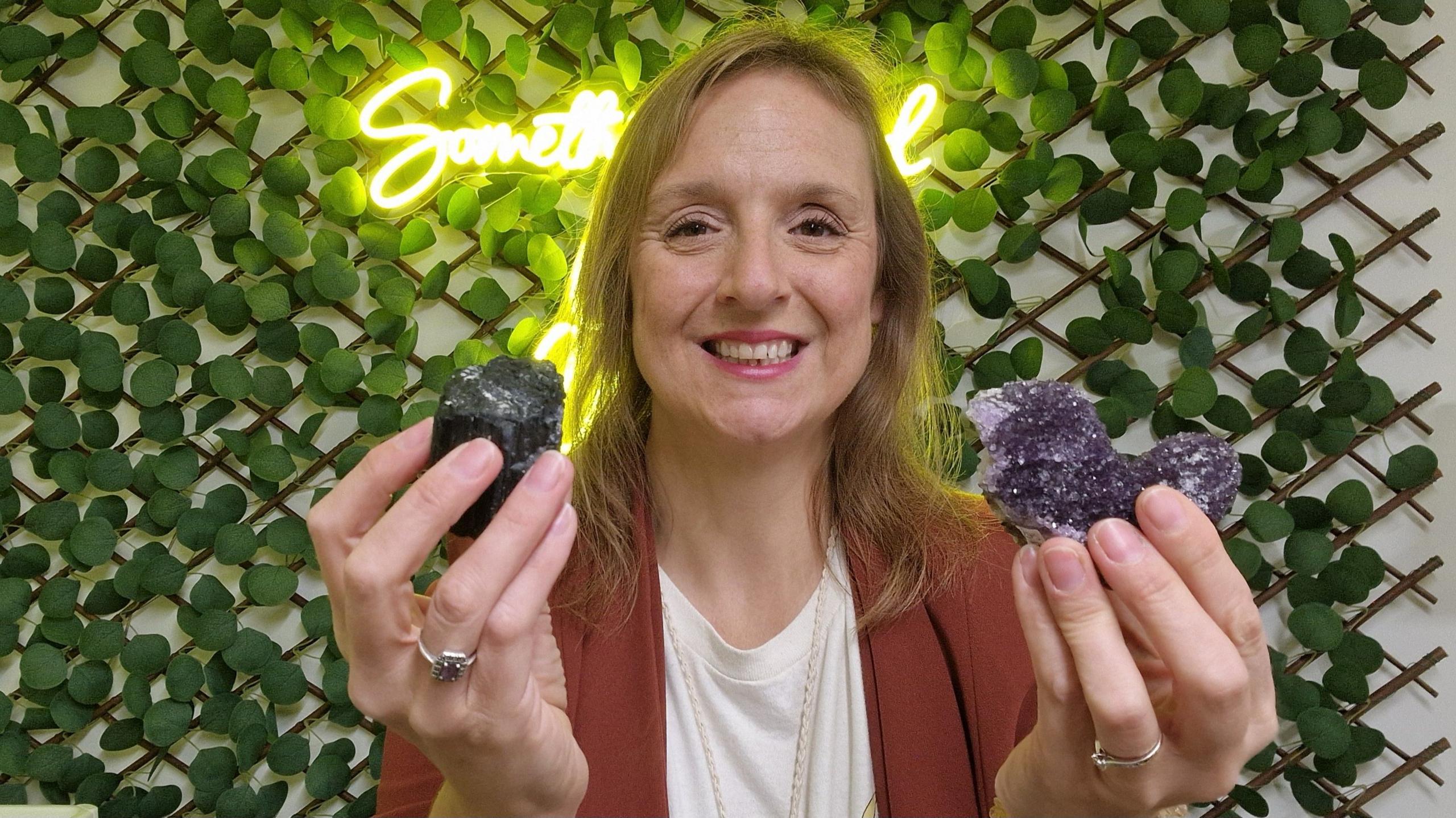 A woman with blonde hair is pictured. She is wearing an amber-coloured jacket with a white top underneath and she is holding two crystals (one is purple and one is black) and smiling at the camera. In the background there is a white wall with a trellis on it that is covered in green leaves. There is also a white light neon sign on the wall.