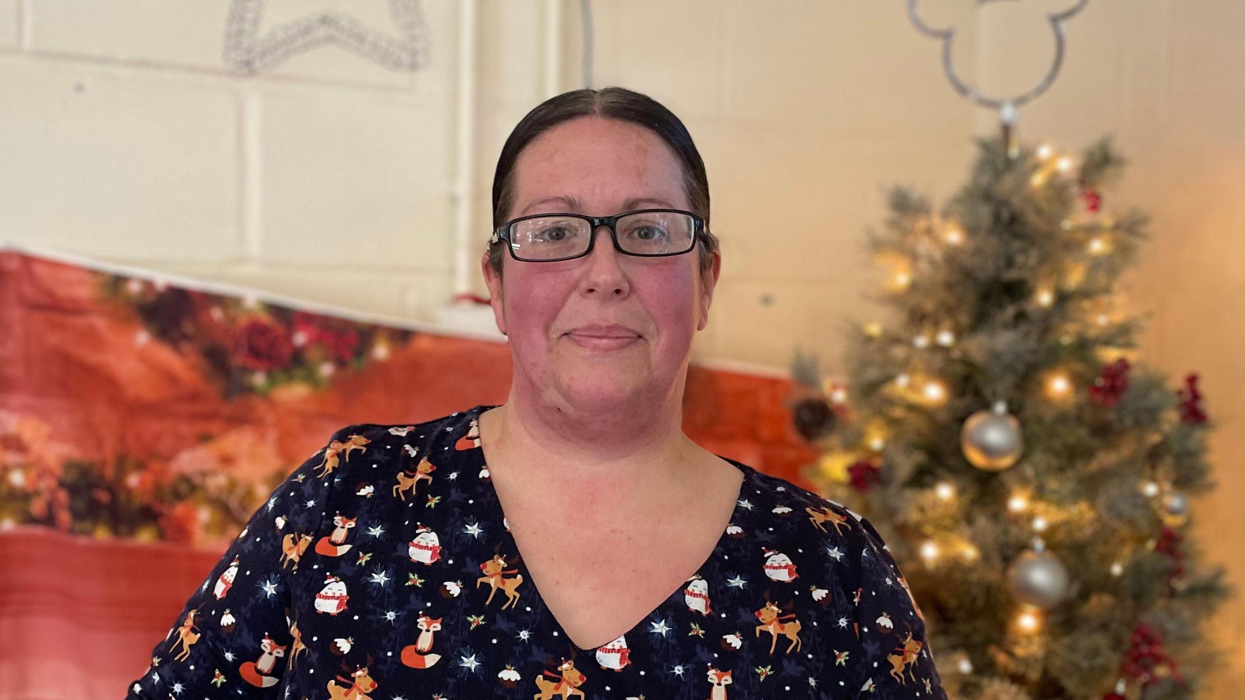 Teresa Duncombe smiling into the camera. She has brunette hair and glasses and has a festive top on. She is standing in front of a Christmas tree.