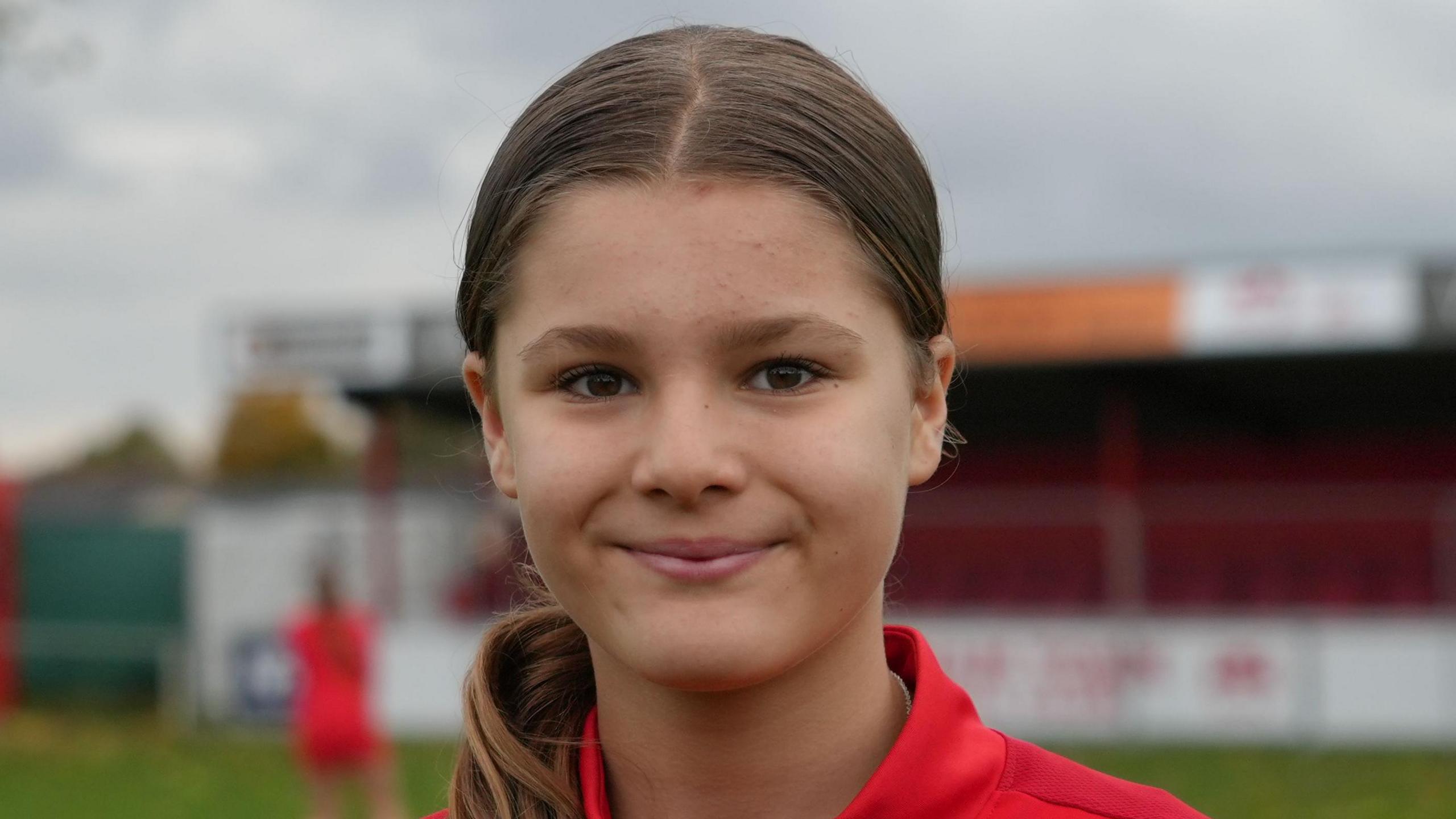 Isabella faces the camera smiling while standing on a football pitch. She is wearing a red jumper with a collar and her long brown hair has been tied up.