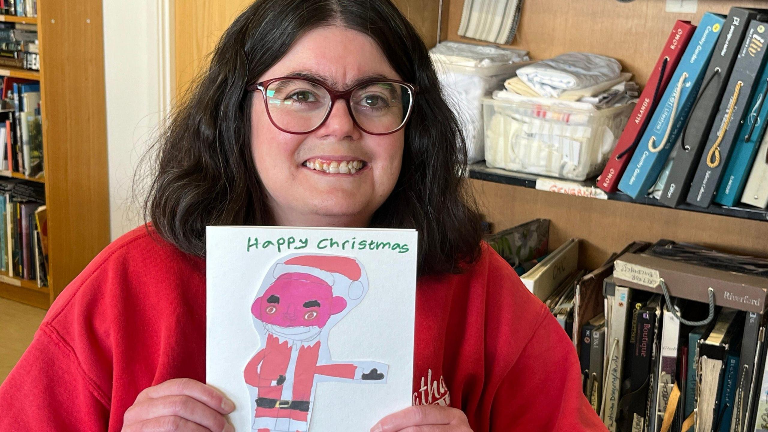 A woman dressed in red holds up a card featuring Father Christmas