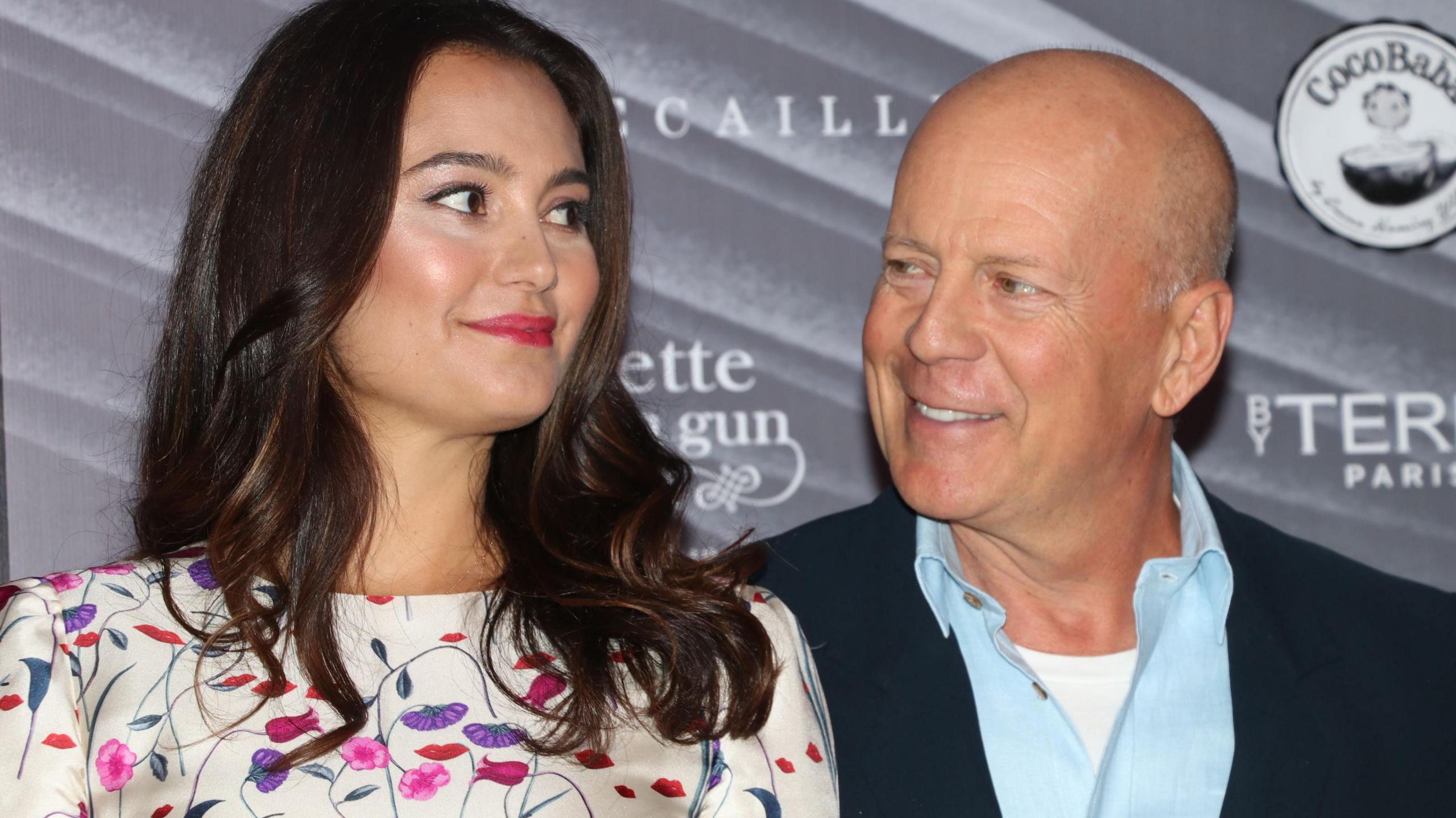 Emma Heming Willis and Bruce Willis (Emma Heming is wearing a flowery dress and has long brown hair, Willis is wearing a light blue shirt and navy jacket)