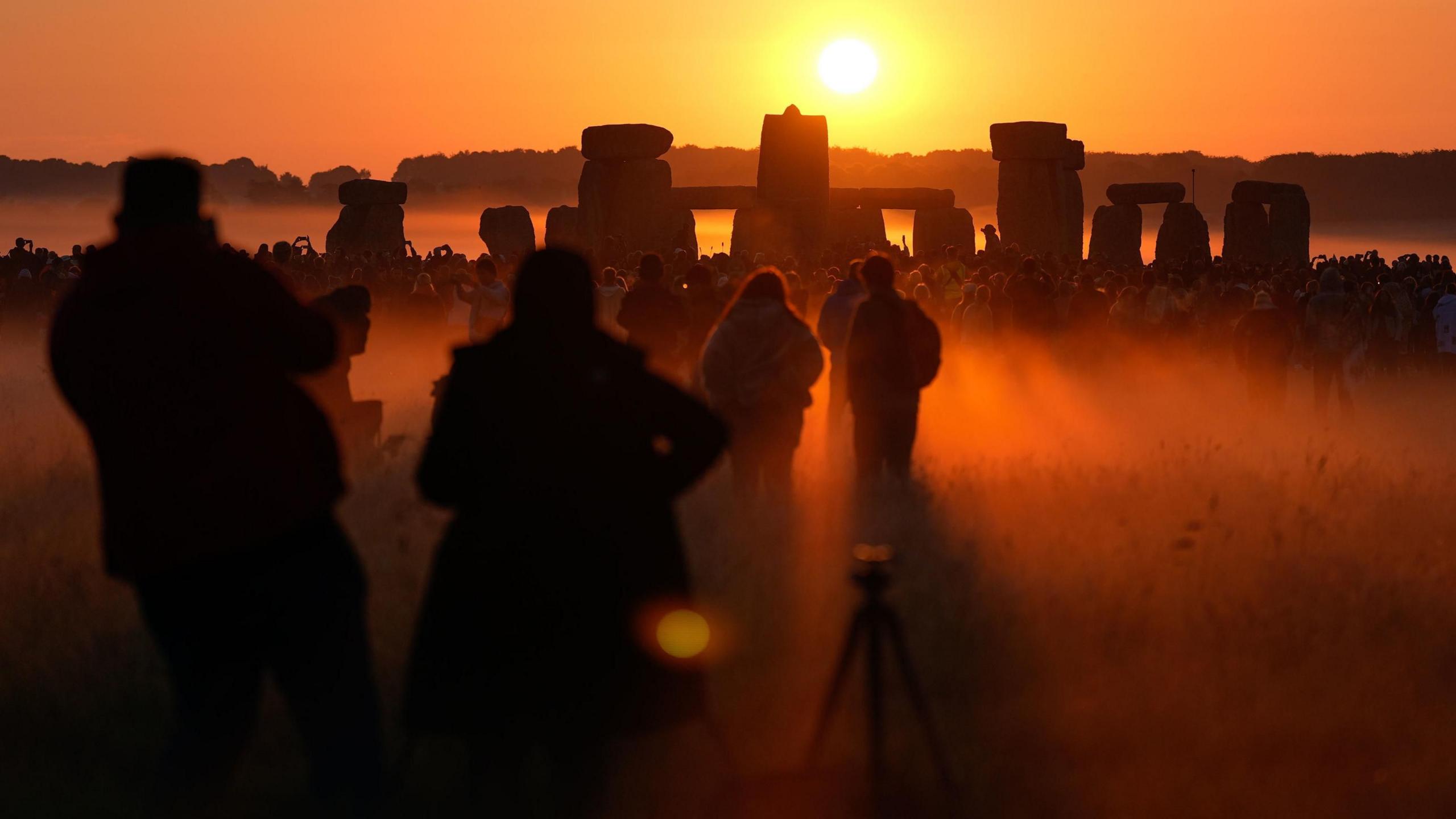 People stand in front of the stones at Stonehenge with the sun rising directly in front of them.