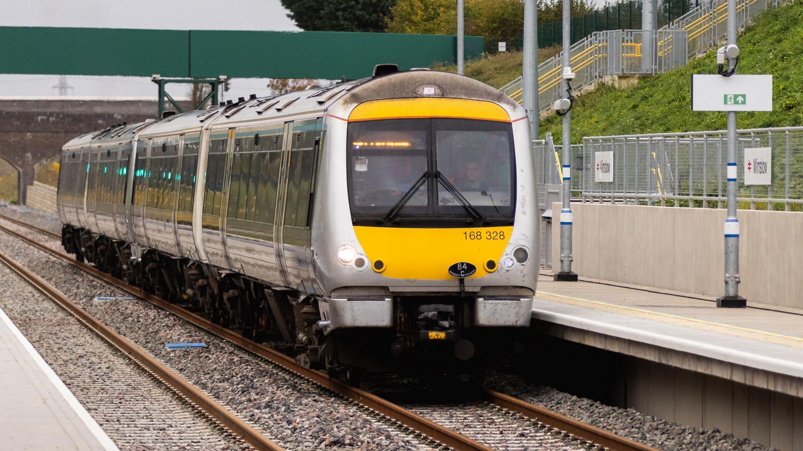A yellow and silver multiple unit train stationary at the platform at Winslow station. There is a green-sided bridge above the platforms behind the train. A grass bank rises to the right alongside the platform.