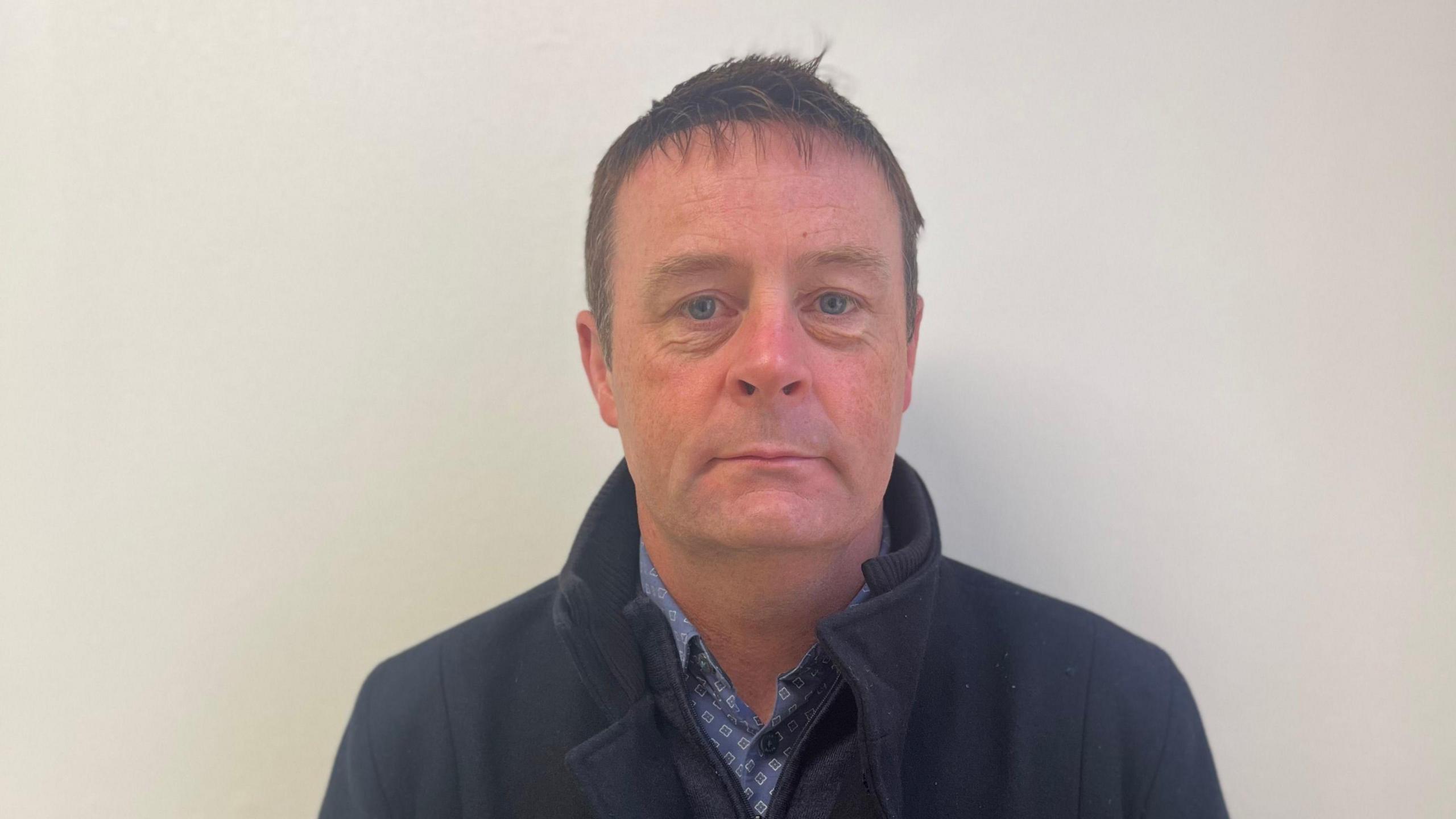 Raymond Gormley, Head of Energy Policy, at the Northern Ireland Consumer Council standing against a white wall. He has a serious but relaxed expression and is wearing a navy coat and blue shirt. His hair is short and brown. 