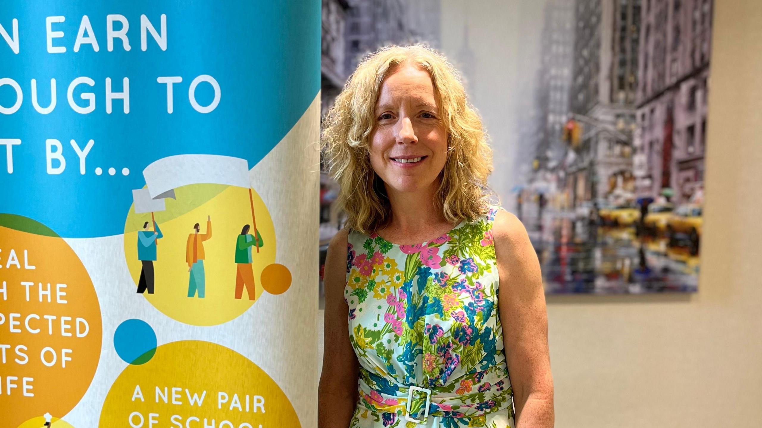  Mary McManus, who has shoulder-length blonde hair, in a floral dress stands next to a pull-up banner with cartoon drawings