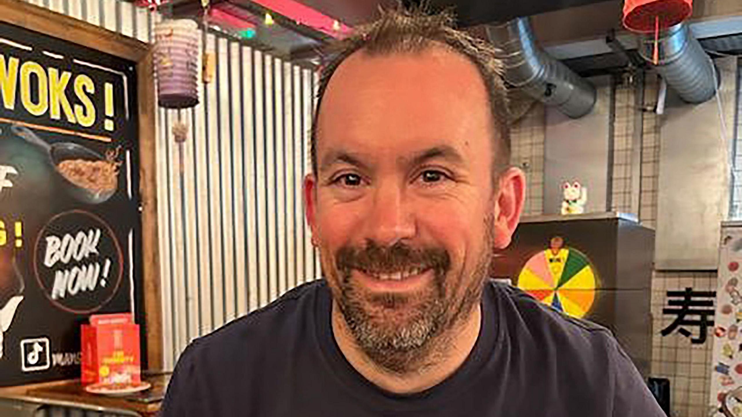 A man with short brown hair and a brown-grey beard sitting in a Chinese restaurant. He is smiling at the camera.