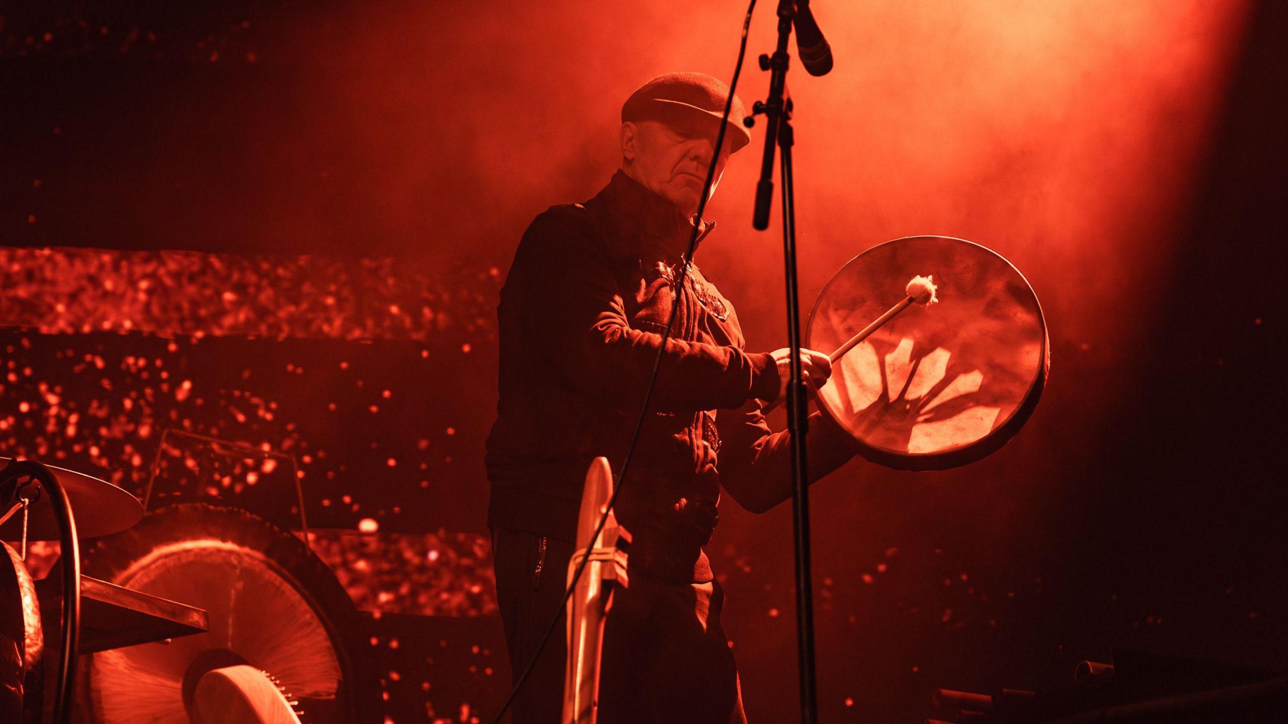 A musician is standing on stage by a microphone. He is playing a hand-held drum.