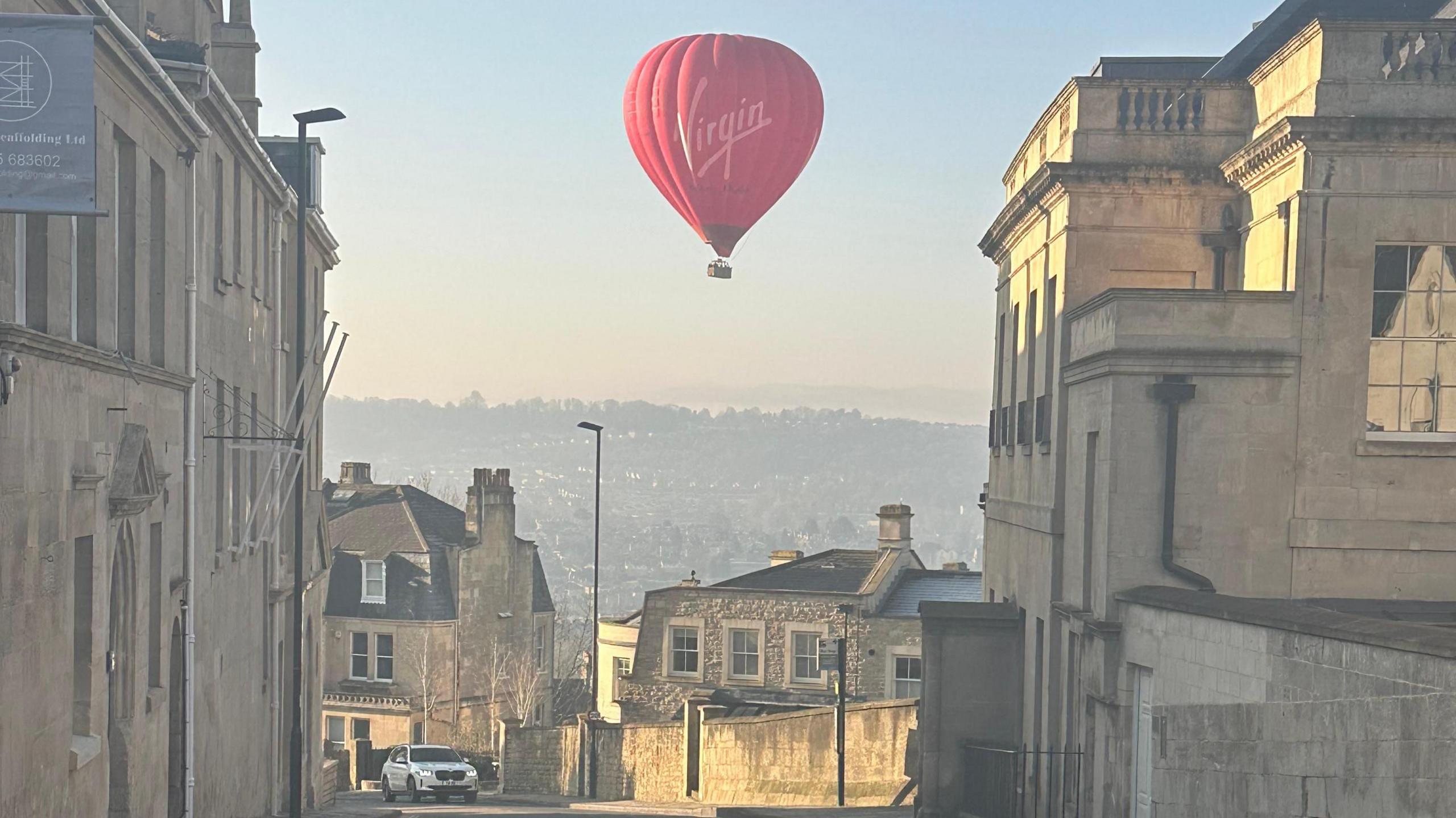 A red hot hair ballon is flying over Charlcombe on a sunny morning. The viewer can see across to Bath over the hill.