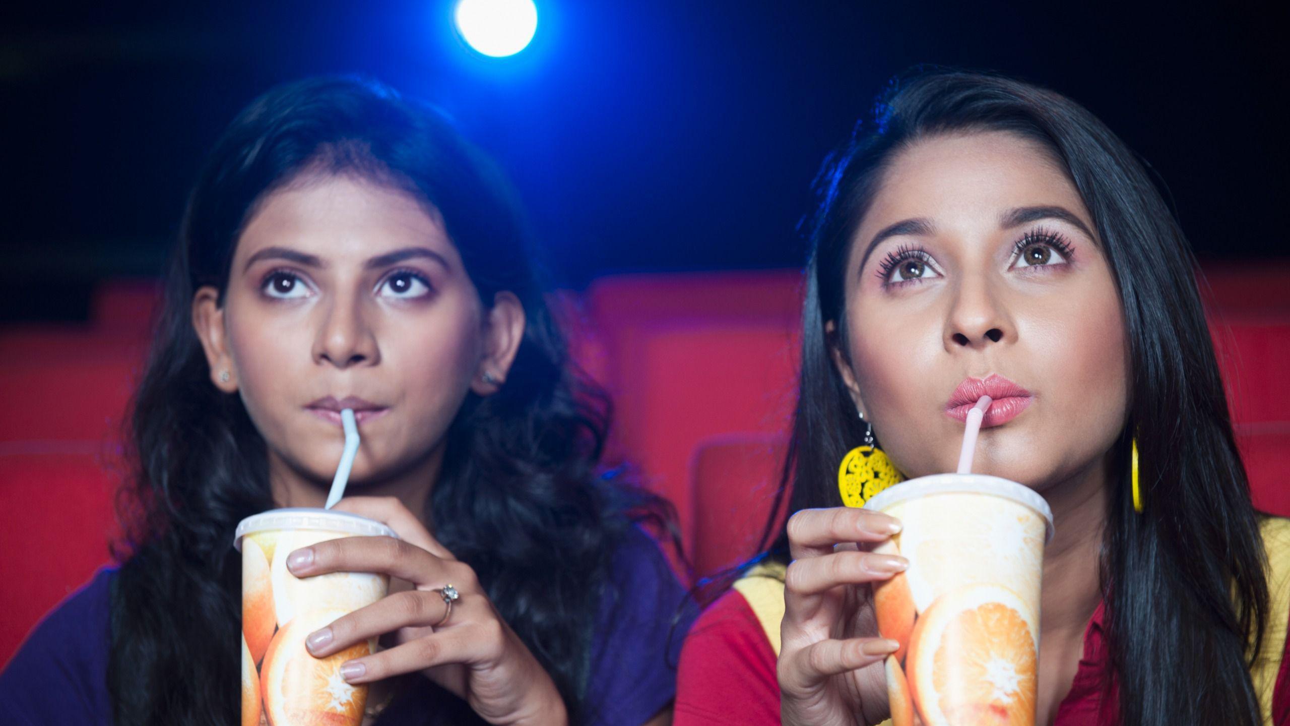 Two women having soft drinks while watching movie in a cinema hall