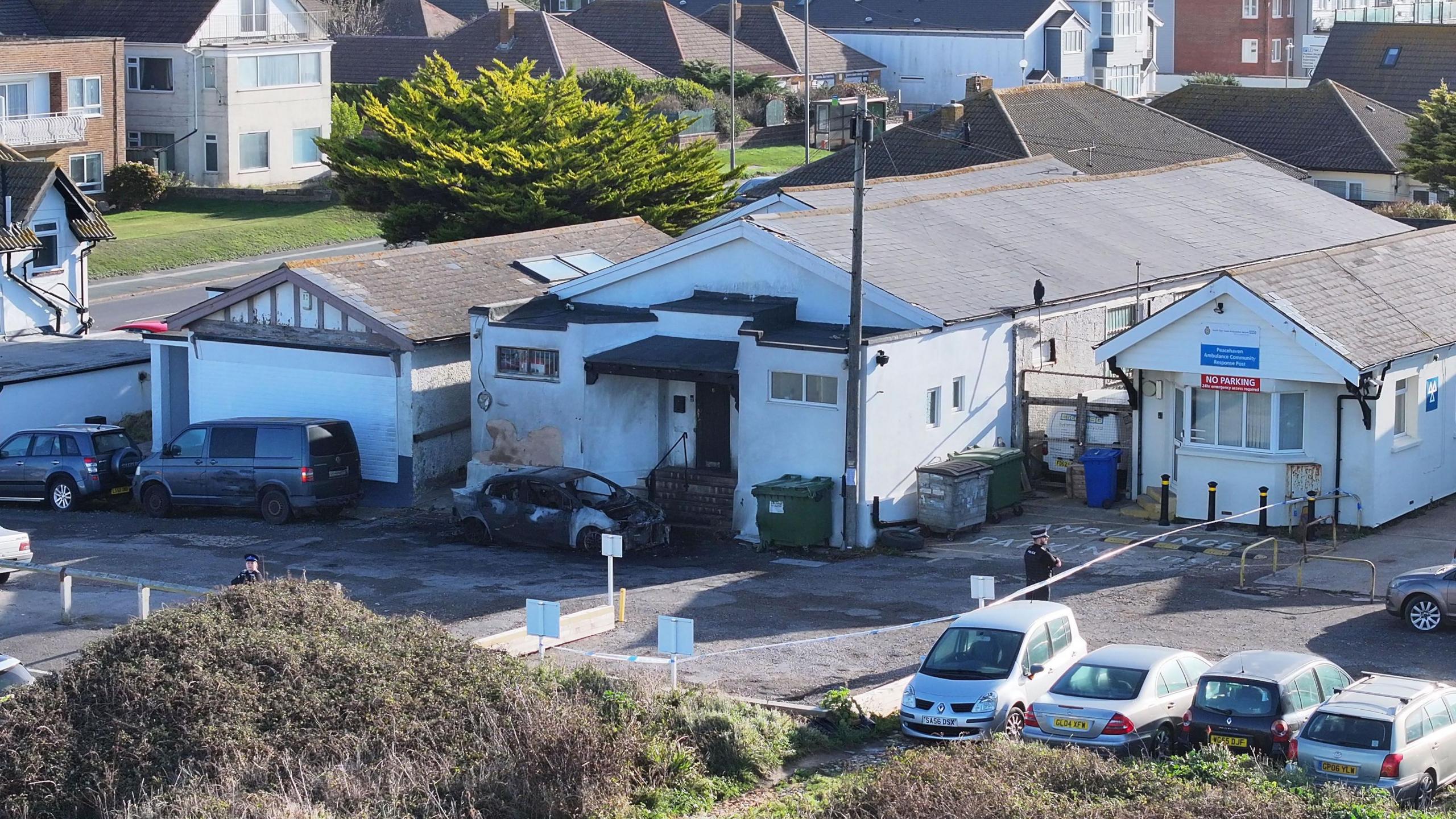 A fire-damaged white building in a coastal English town. A burnt-out car is parked in front of the mosque entrance. There is police tape around the scene and police officers standing guard.