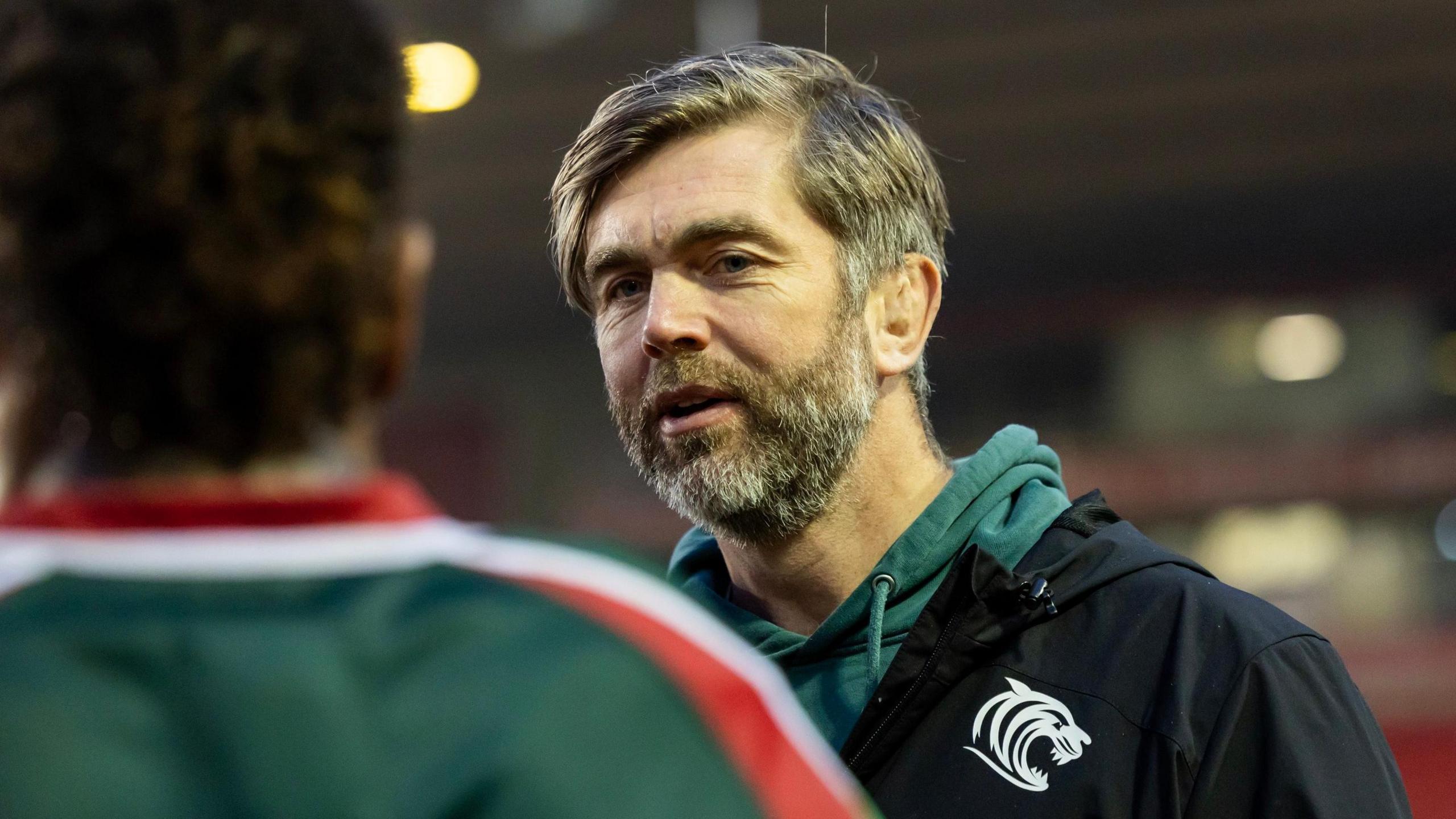 Geoff Parling talking to one of his Leicester Tigers players and he is wearing a green hoodie and black weather proof coat with the Tigers emblem on it.