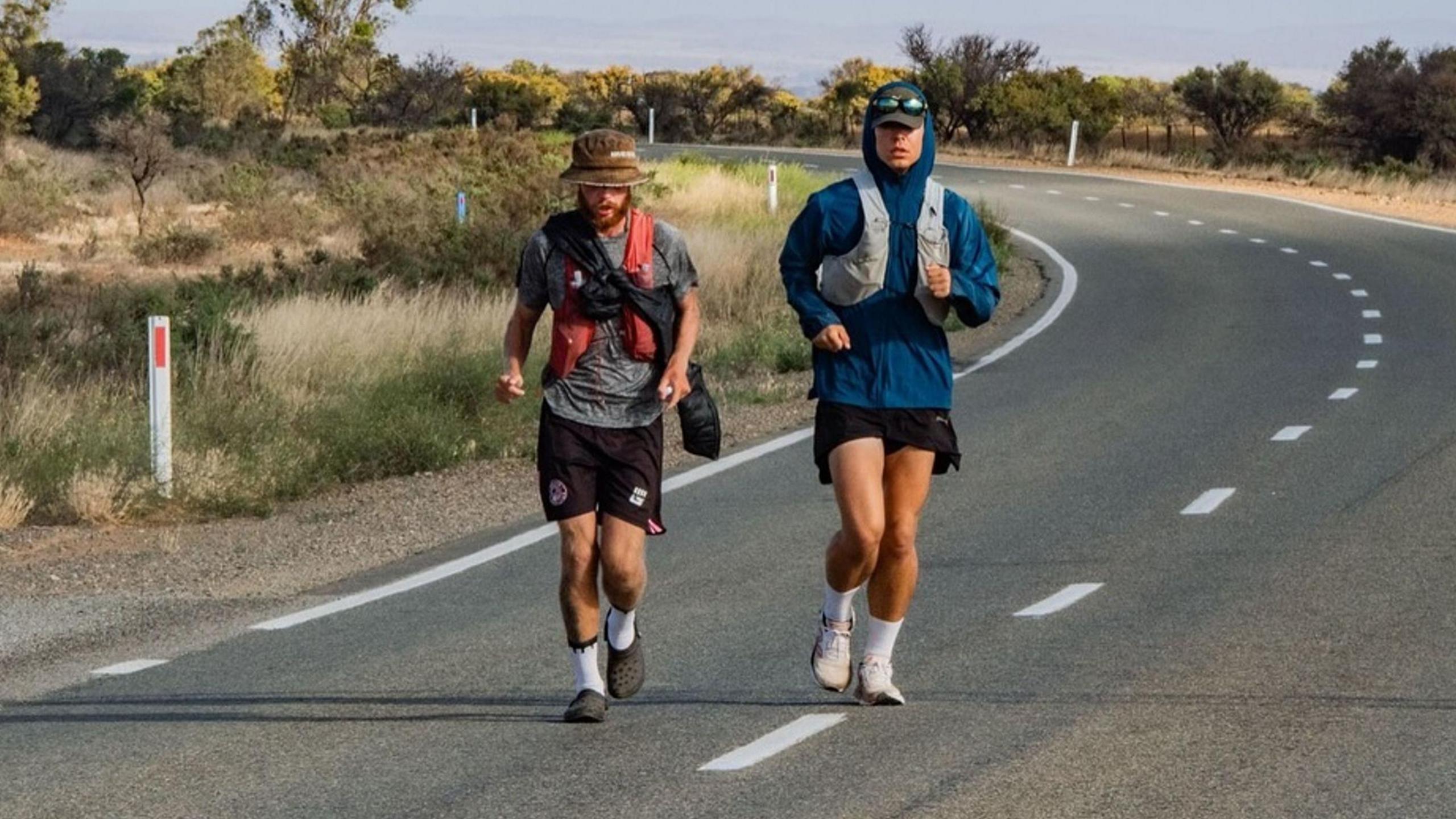 Jack Pitcher and Joshua Smith running along a road, running along the centre of the road. Jack Pitcher is wearing Crocs shoes, shorts, grey T-shirt and a brown hat. Joshua Smith is wearing trainers, black shorts and a blue hooded top with sunglasses resting on the top of a cap