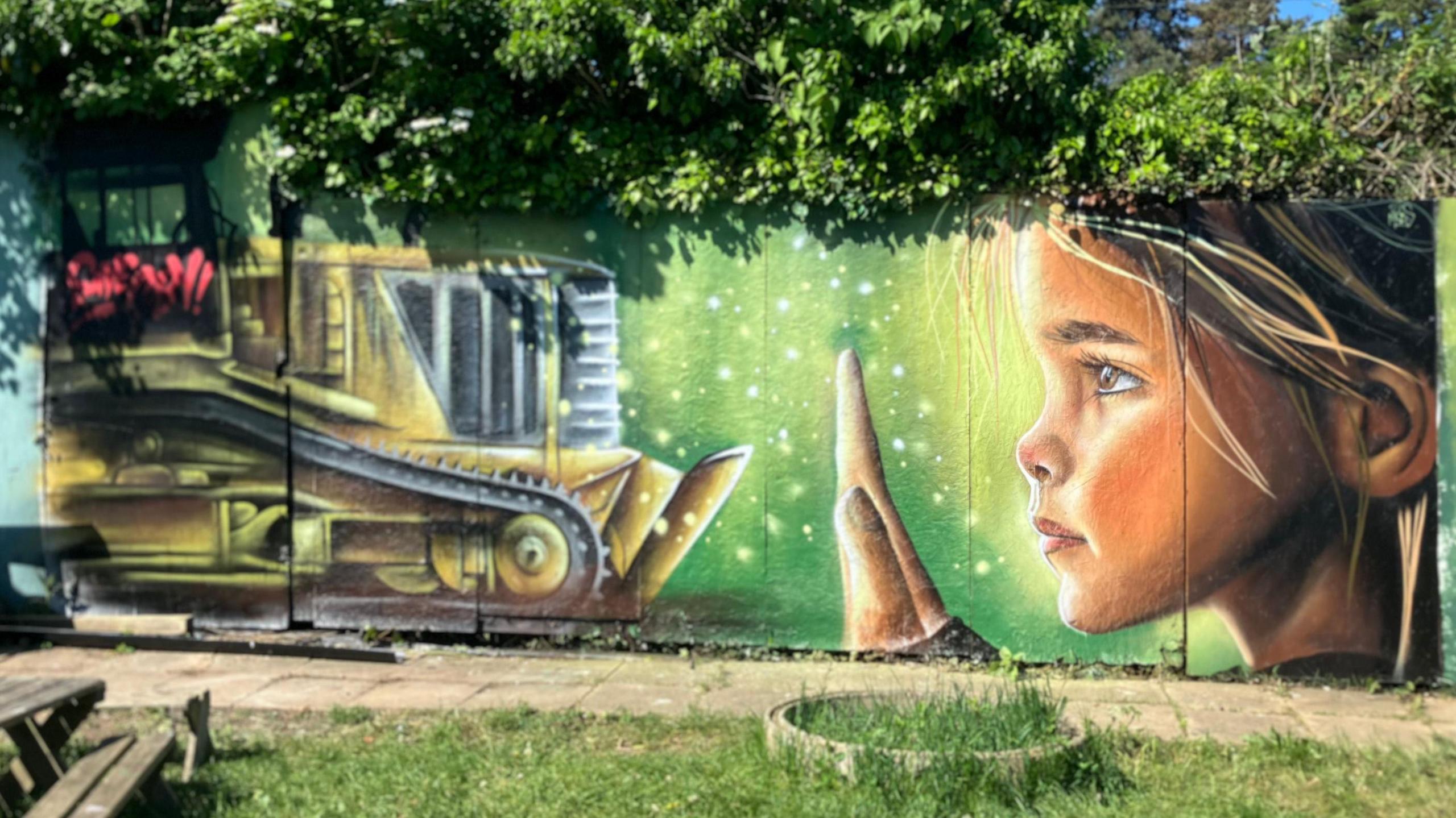 Mural of a girl holding up a hand to a yellow bulldozer- with a green background painted on a wall lined with trees