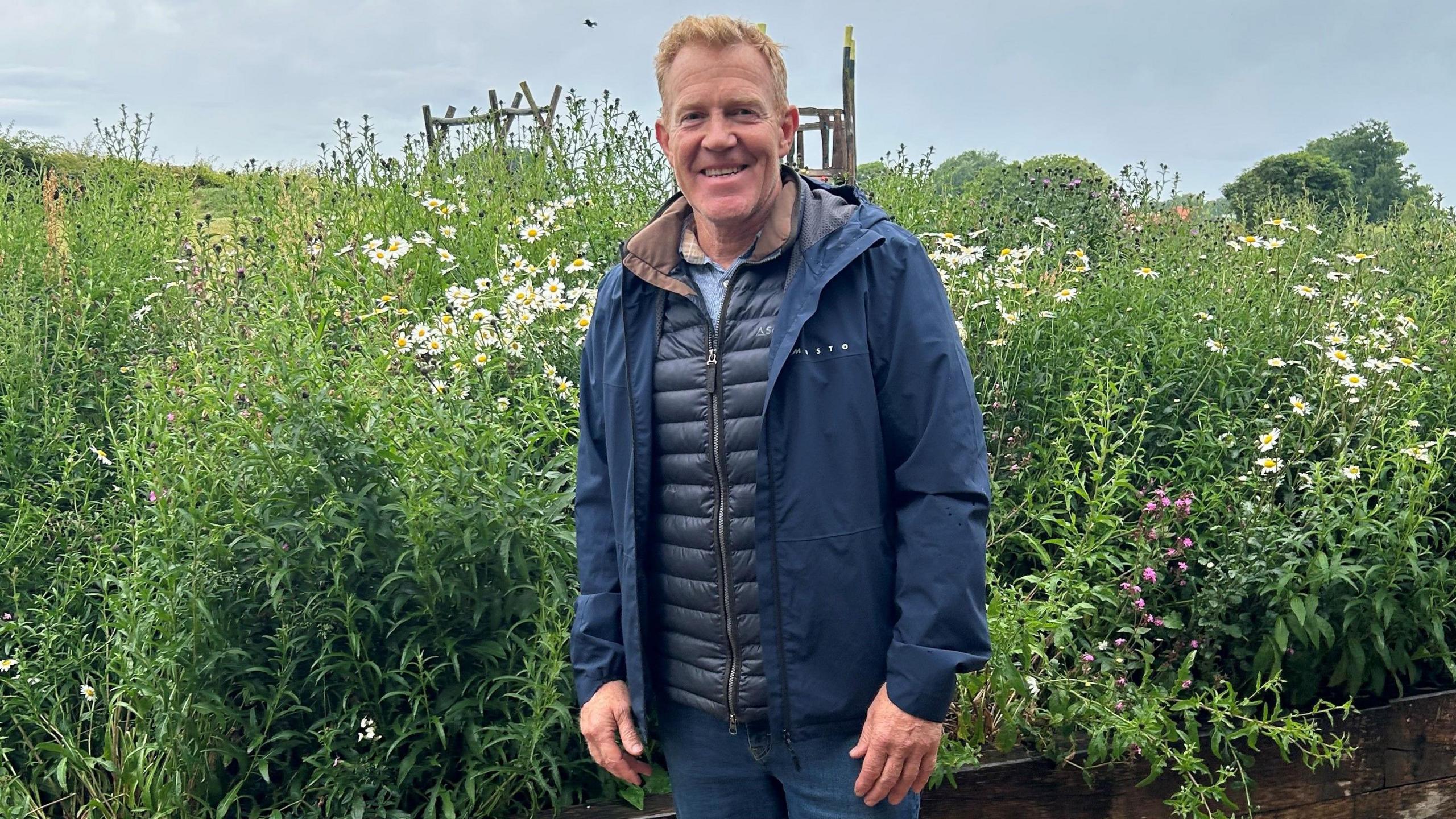 Adam Henson, wearing all navy blue - blue jeans and a blue waterproof - stands in front of a tall bed of wildflowers