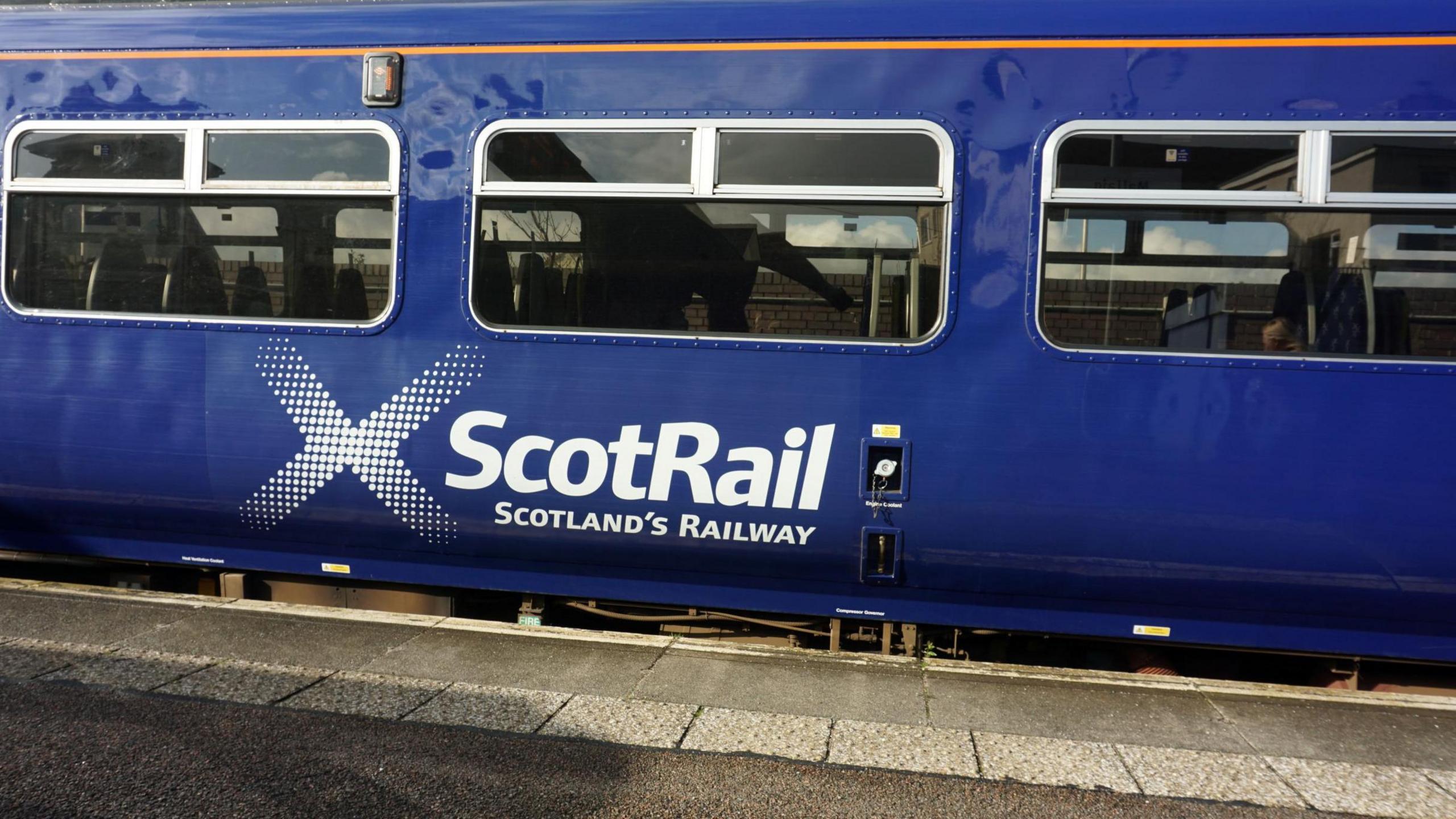 A carriage on a ScotRail train at a station platform. The carriage is blue with the words "ScotRail" and "Scotland's Railway".