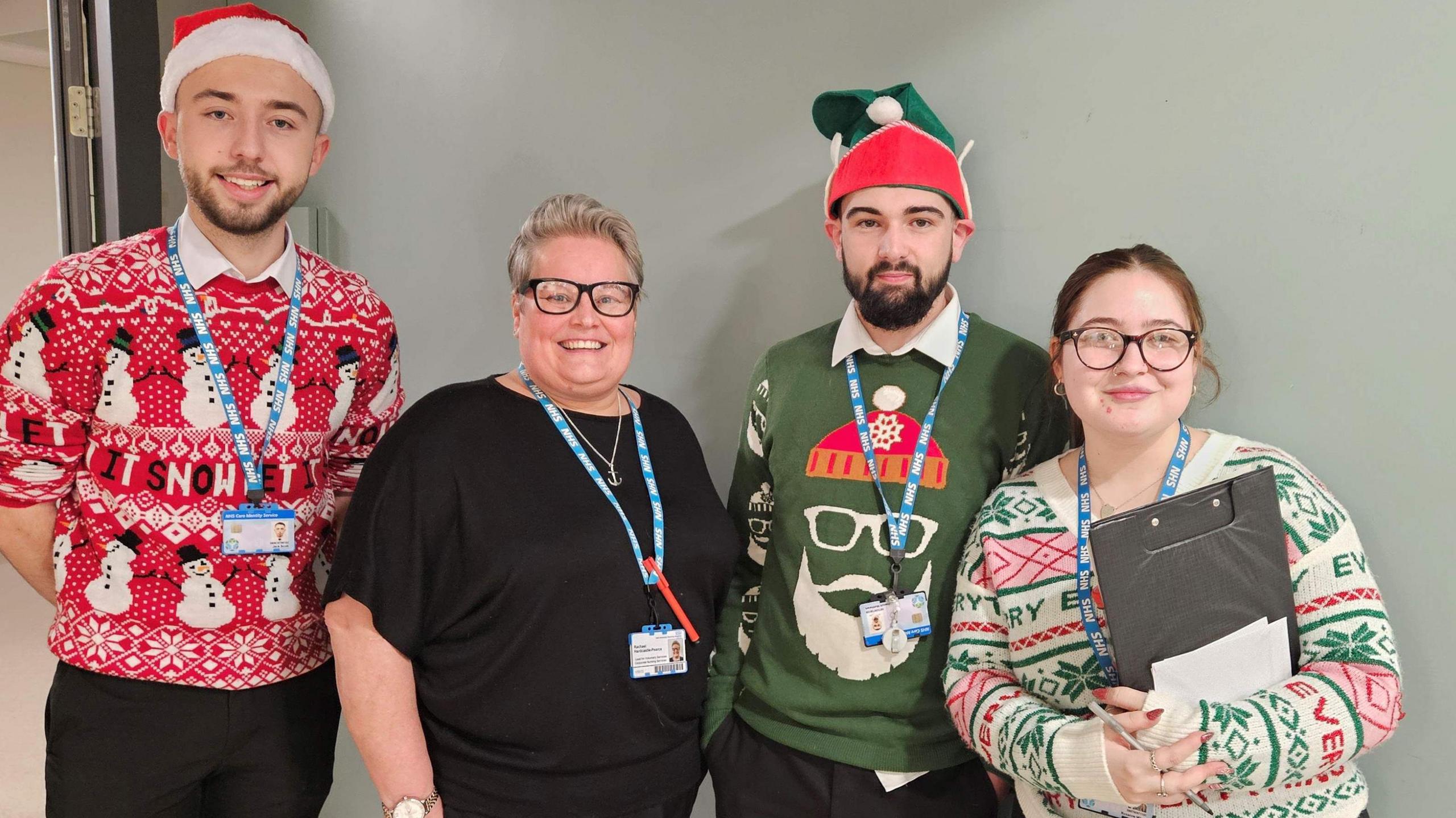 Two men and two women are dressed in Christmas jumpers and are smiling for the camera.  They have hospital lanyards on and one of the women is holding a clip board. The two men are wearing Santa hats.