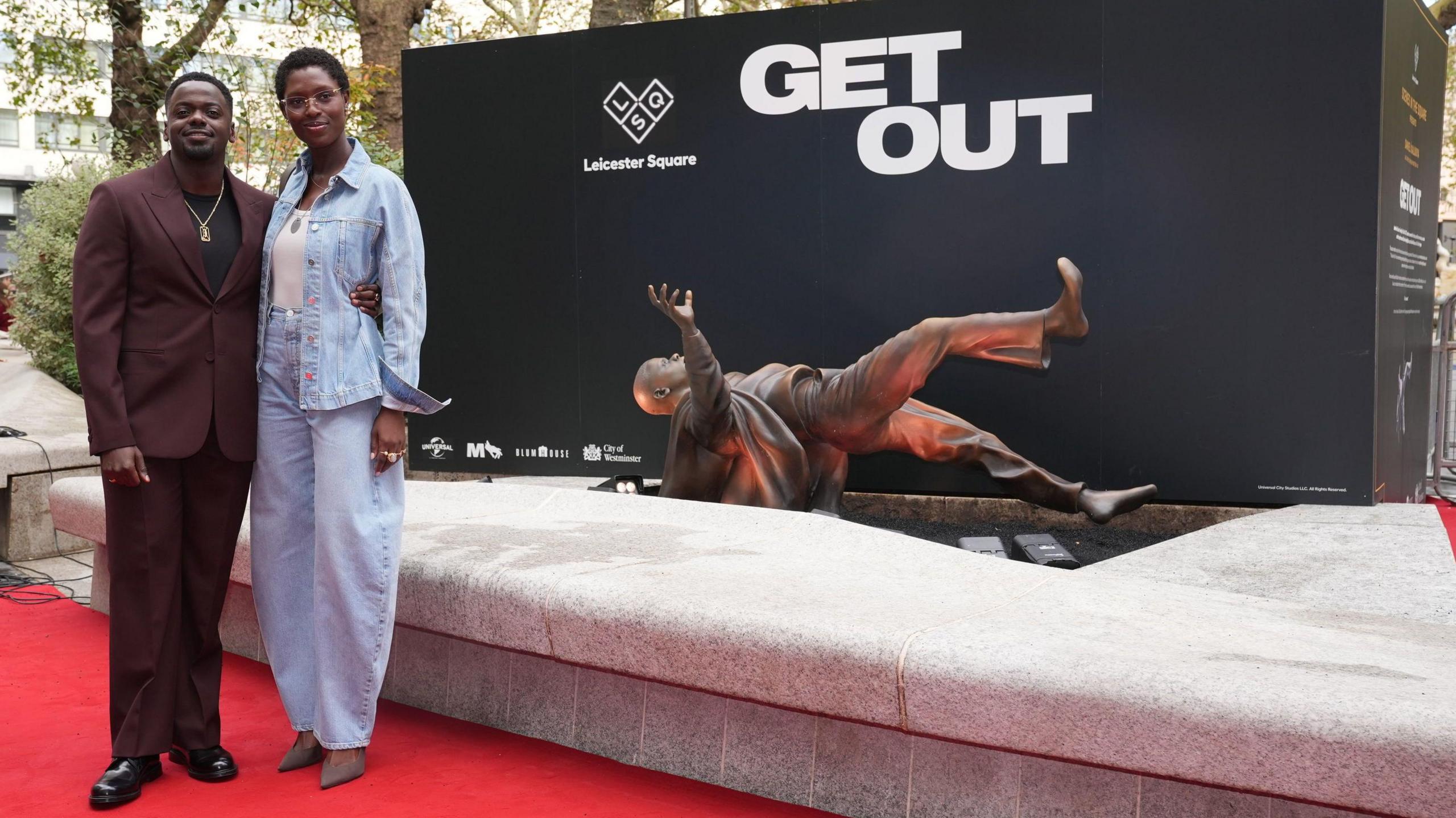 Jodie Turner-Smith and Daniel Kaluuya pose on a red carpet beside the horizontal statue of Mr Kaluuya as his character in Get Out. Mr Kaluuya is dressed in a mauve suit and Ms Turner-Smith is wearing a blue denim jacket and trousers. 