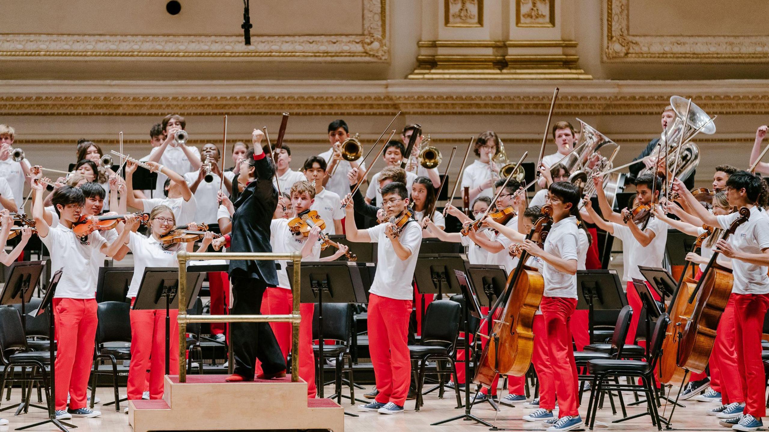 The National Youth Orchestra 2 onstage - their conductor is conducting them with a flourish while the orchestra members are in white shirts and red trousers. 