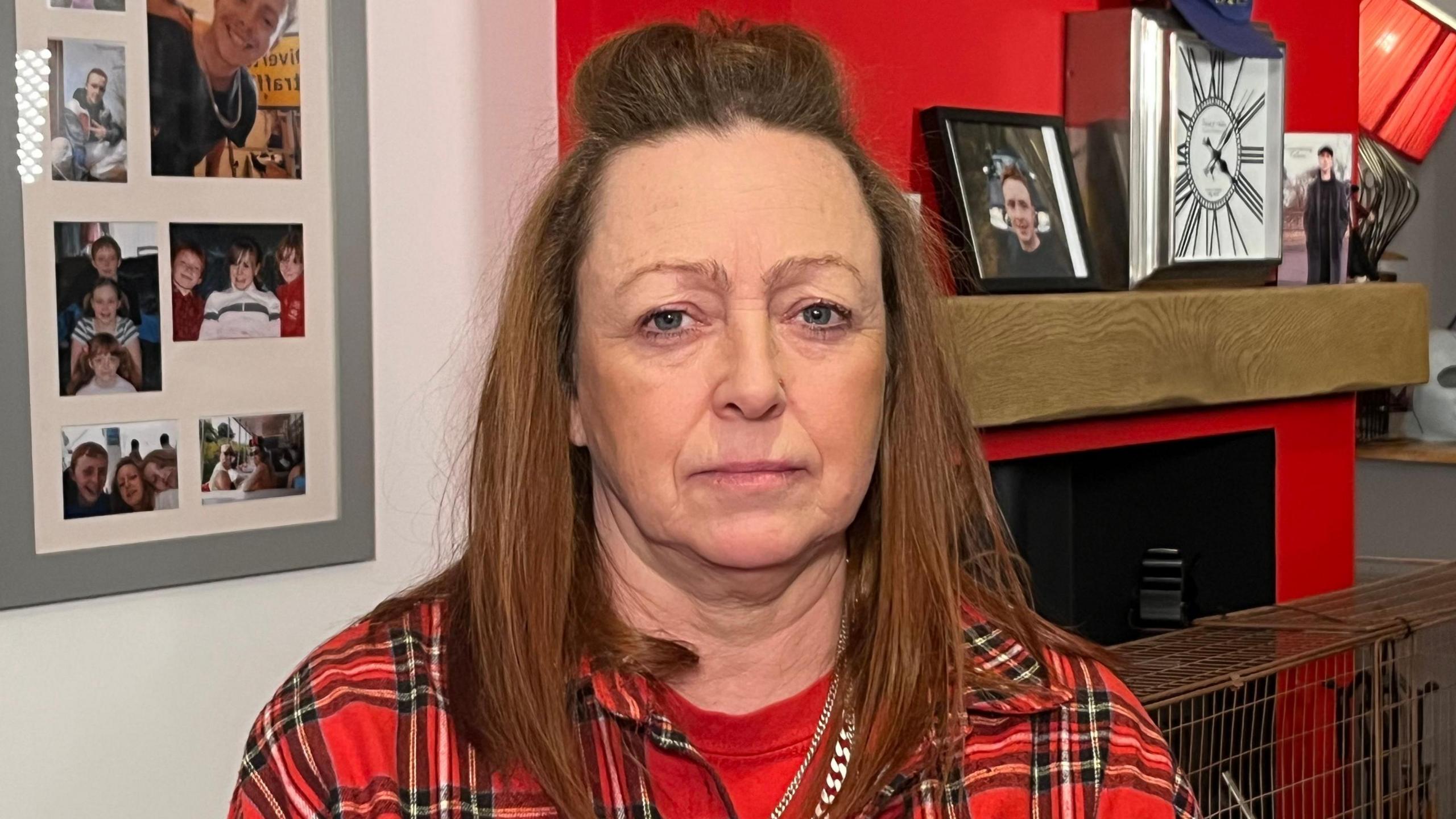 Jillie Slope, wearing a tartan shirt and staring at the camera. Behind her are images and items honouring her son on the mantlepiece of their fire place and on the walls. 