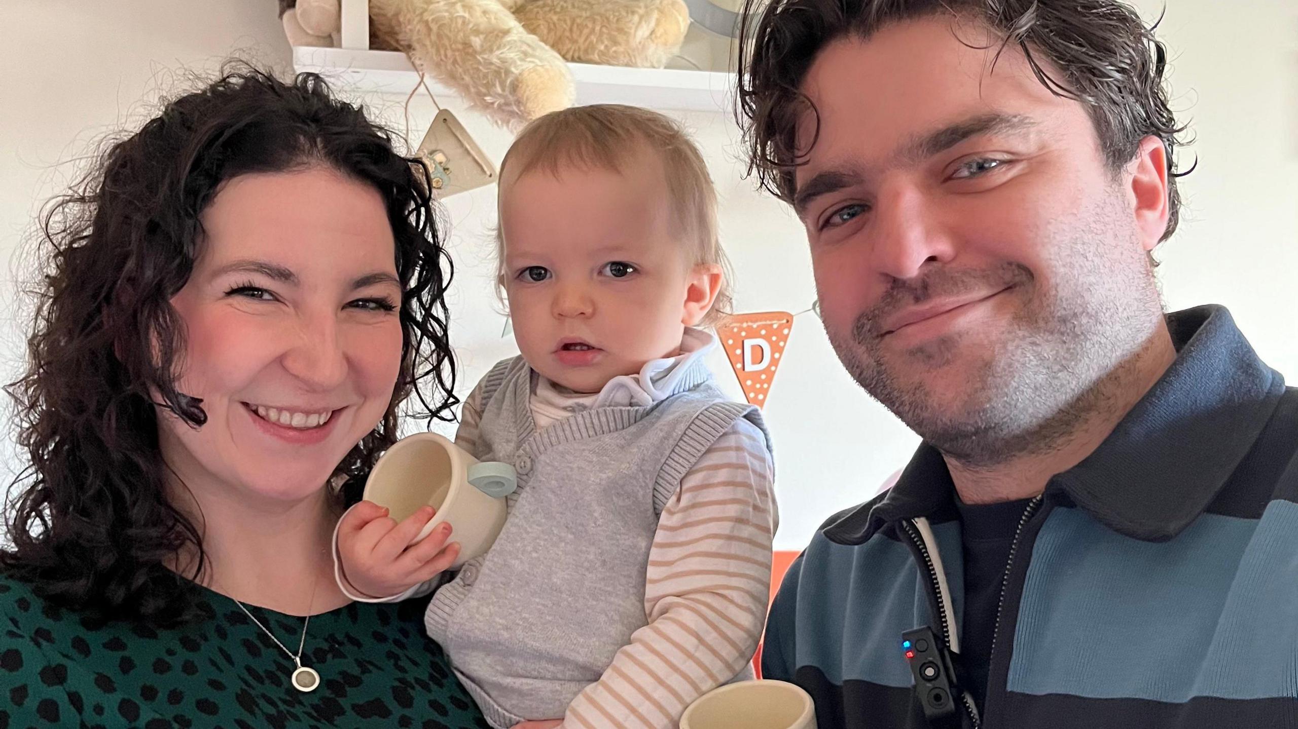 Family portrait of Naomi, Roddy and Ali. The parents are smiling at the camera while baby Roddy stares with a questioning expression. The picture appears to be taken in a child's bedroom with a large teddy bear visible behind. Mum has long, black, curly hair and is wearing a green top with black spots. Dad has dark hair and stubble and is wearing a blue sweater with thick stripes.