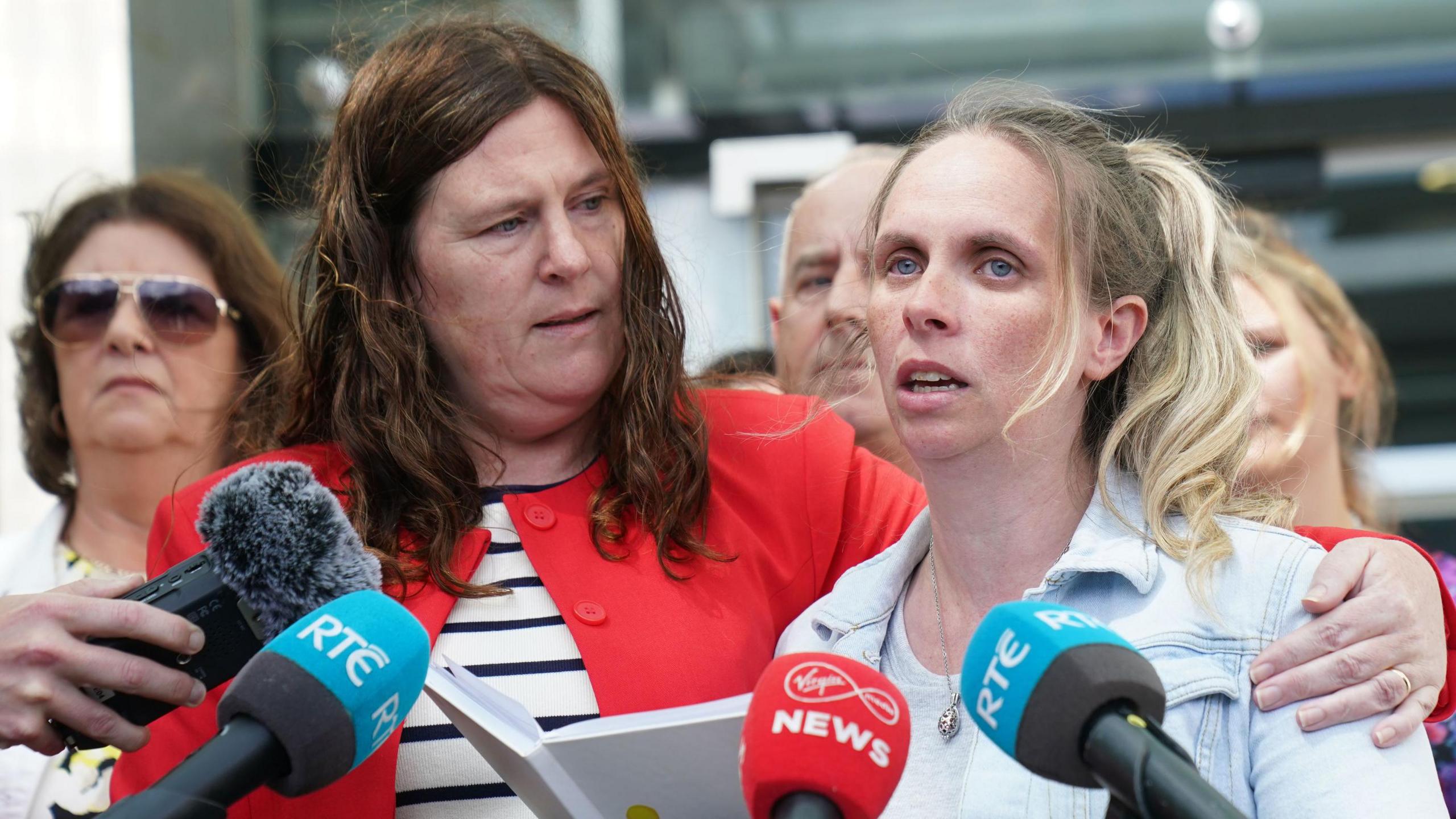 Members of Tina Satchwell's family gathered outside the Central Criminal Court in Dublin to speak to reporters. Sarah Howard speaks into a number of microphones. She has long, blonde hair tied back in a ponytail and is wearing a denim jacket over a white dress. Another of Tina Satchwell's relatives has her arm on Sarah's shoulder